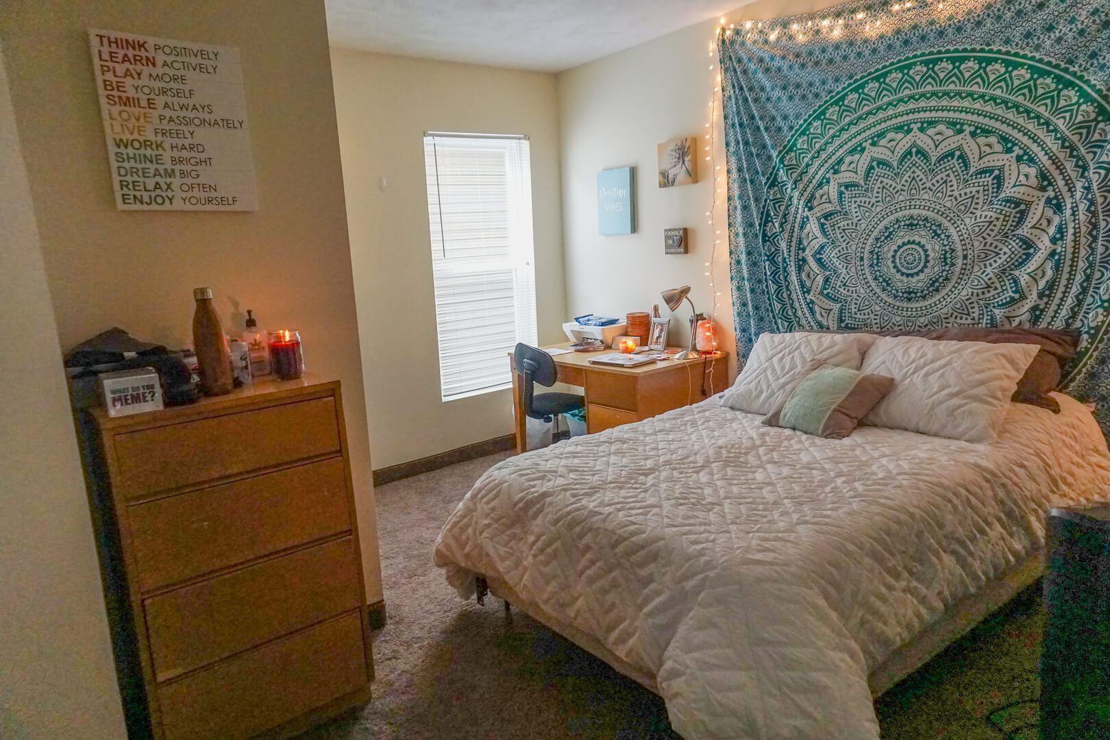 A bedroom with a bed , desk , dresser and a tapestry on the wall.
