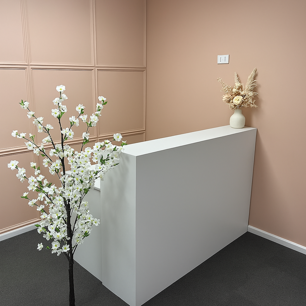 White reception desk in a pink-walled office with a floral arrangement and a small tree.
