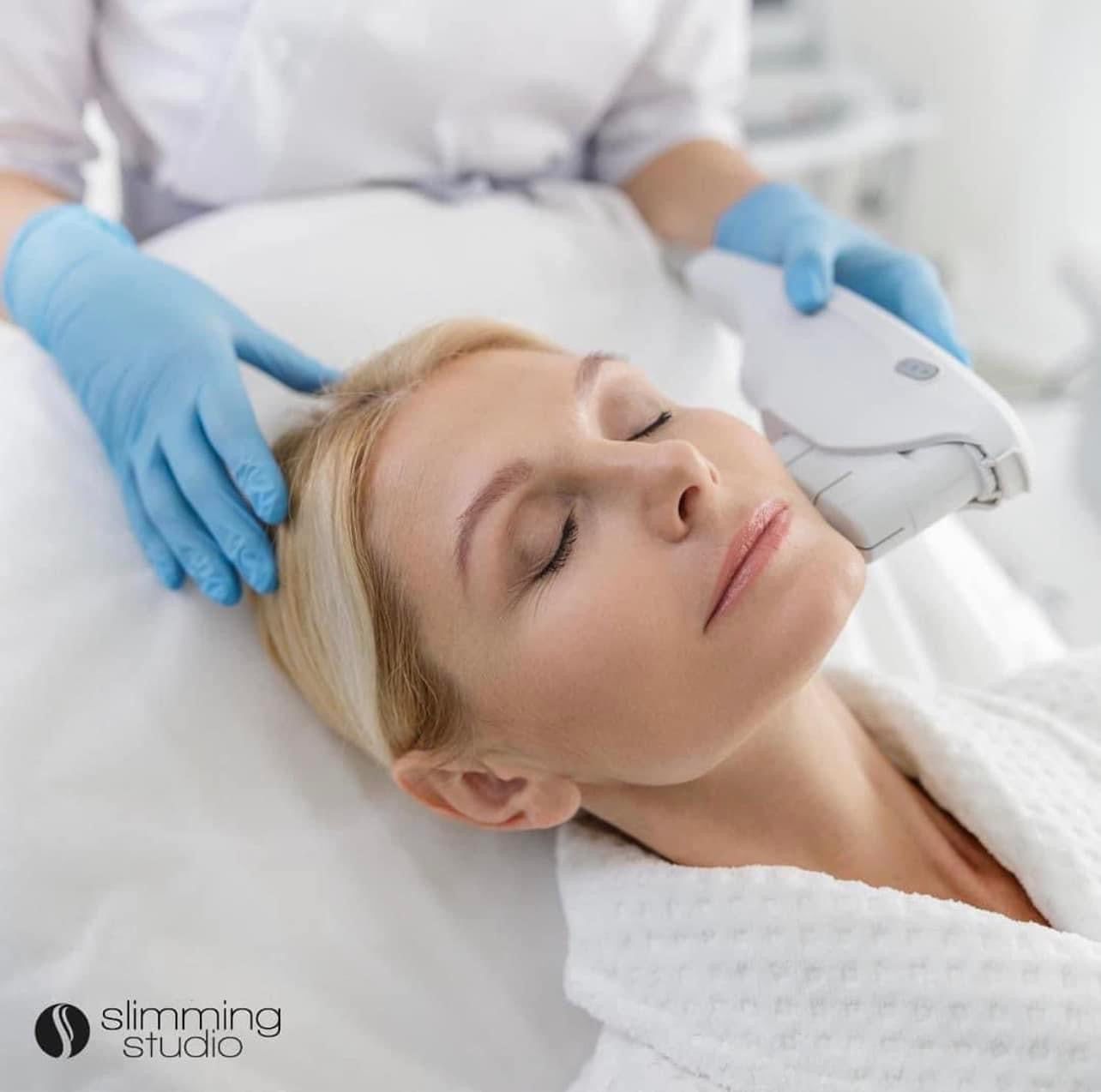 Woman receiving cosmetic facial treatment with a device. White setting; blue gloves.