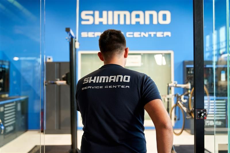 Una persona che indossa una maglietta del Centro Assistenza Shimano si trova in un'area di lavoro blu, con il logo del marchio, e dispone di attrezzature per la riparazione di biciclette.
