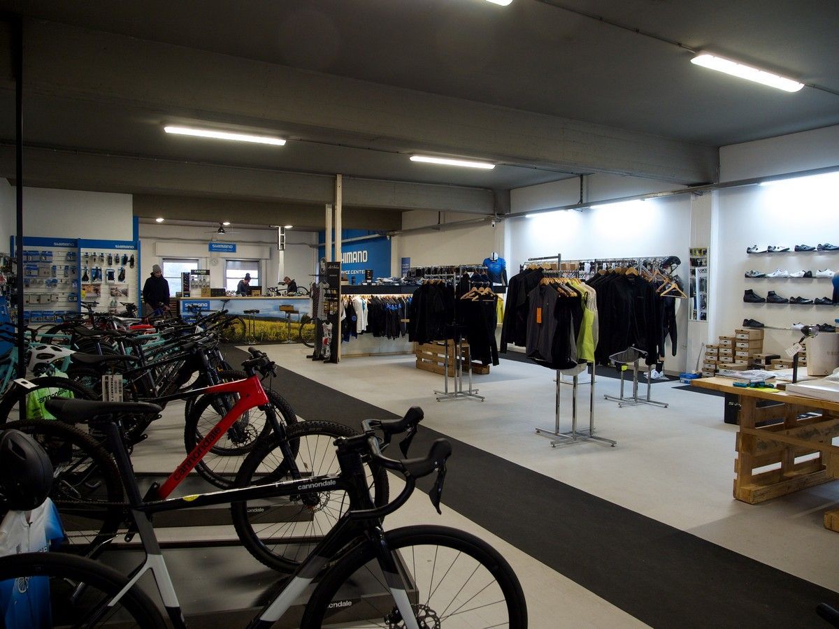 Un negozio di biciclette al dettaglio con file di biciclette in primo piano, appendiabiti al centro e calzature su scaffali a muro.