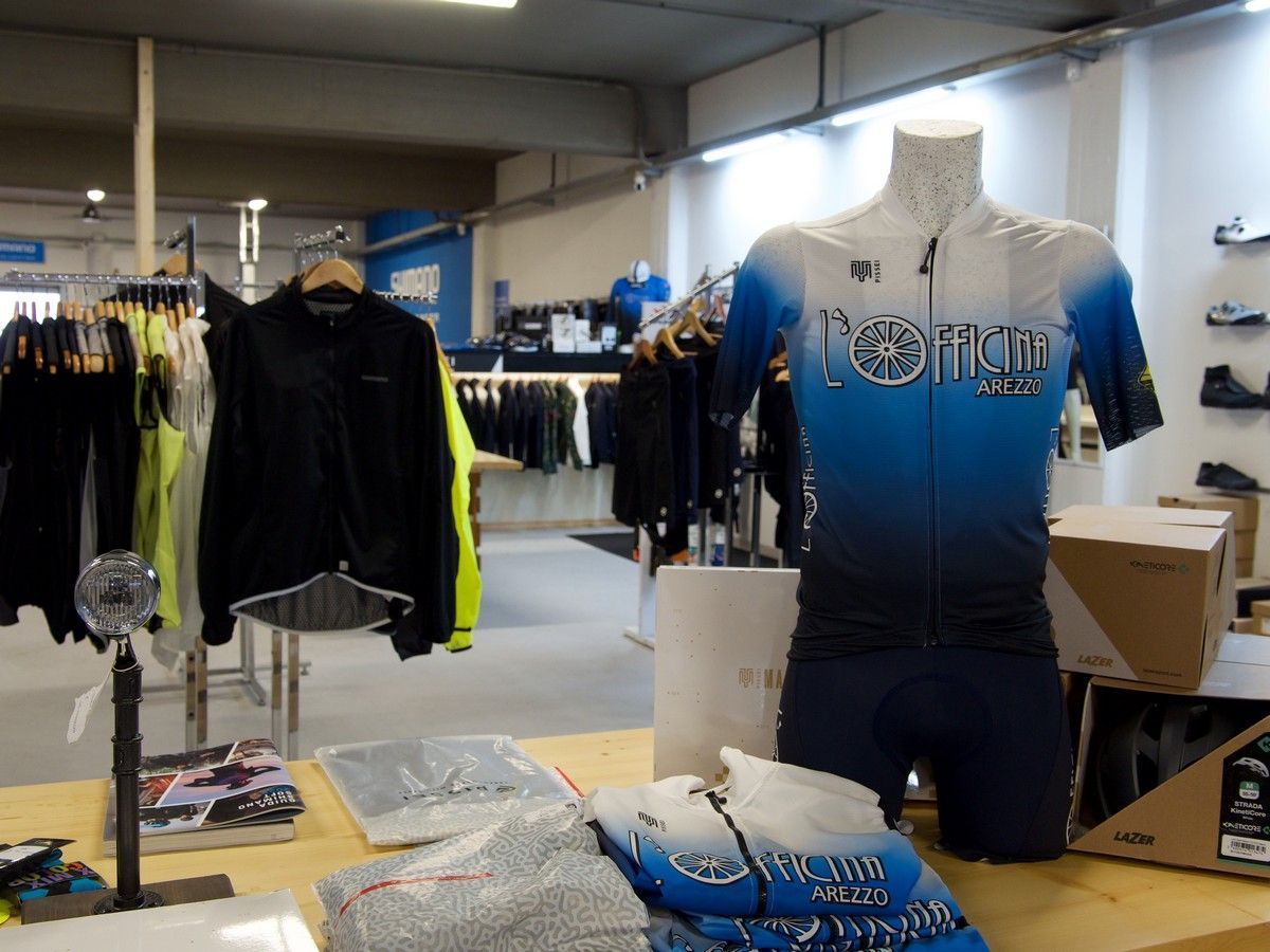 Una maglia da ciclismo blu e bianca su un manichino in un negozio di abbigliamento, con scaffali di vestiti e attrezzature sportive nelle vicinanze.