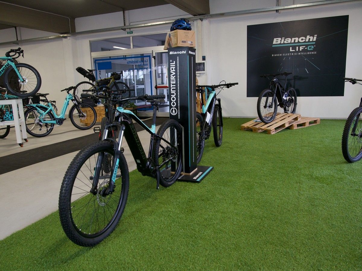 Uno showroom che espone diverse e-mountain bike Bianchi su un prato artificiale, con un espositore a colonna con il logo del marchio al centro.