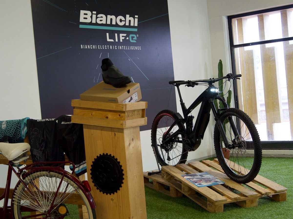 Una bicicletta elettrica Bianchi nera esposta su un pallet di legno in uno showroom, con un'insegna a muro del marchio e una bicicletta d'epoca nelle vicinanze.