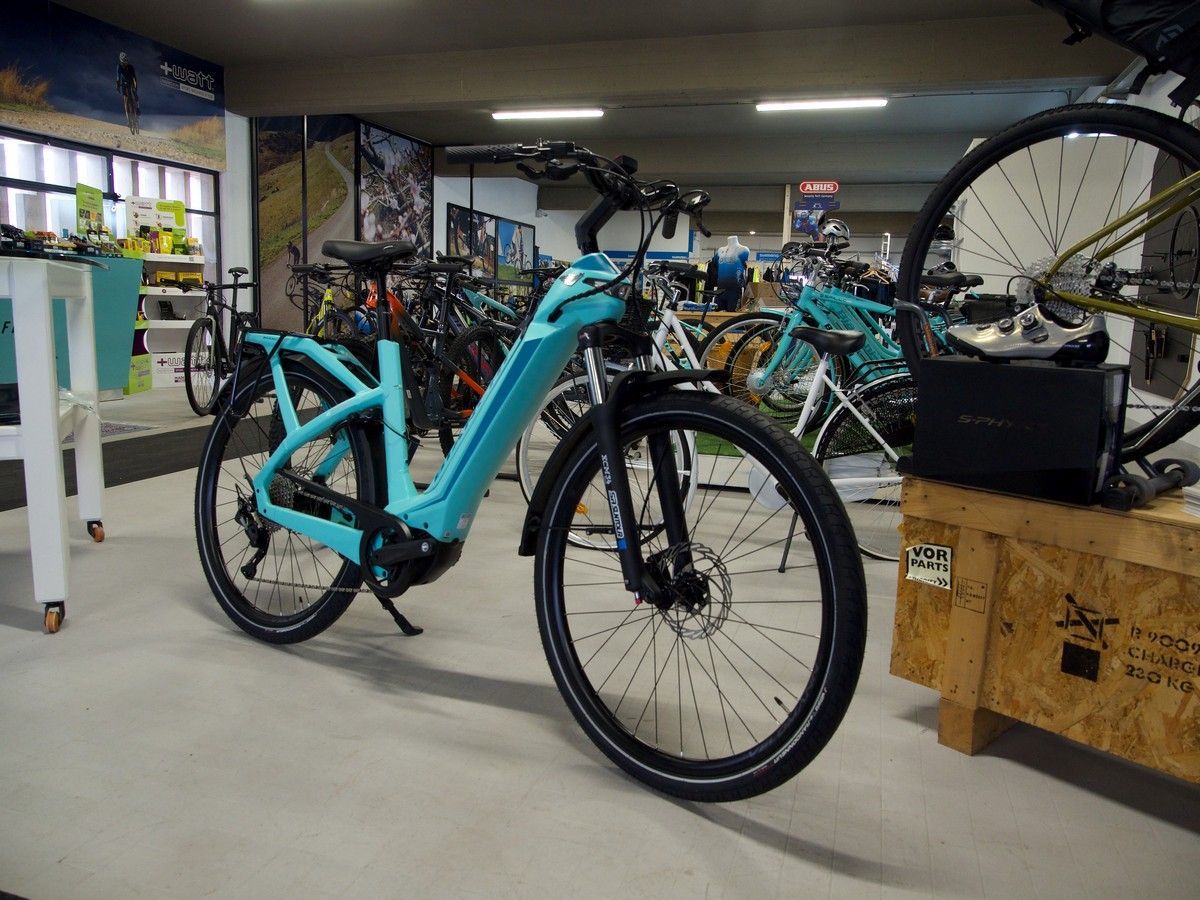 Una bicicletta elettrica da pendolare color verde acqua parcheggiata all'interno di un negozio di biciclette, circondata da altre biciclette e attrezzature.