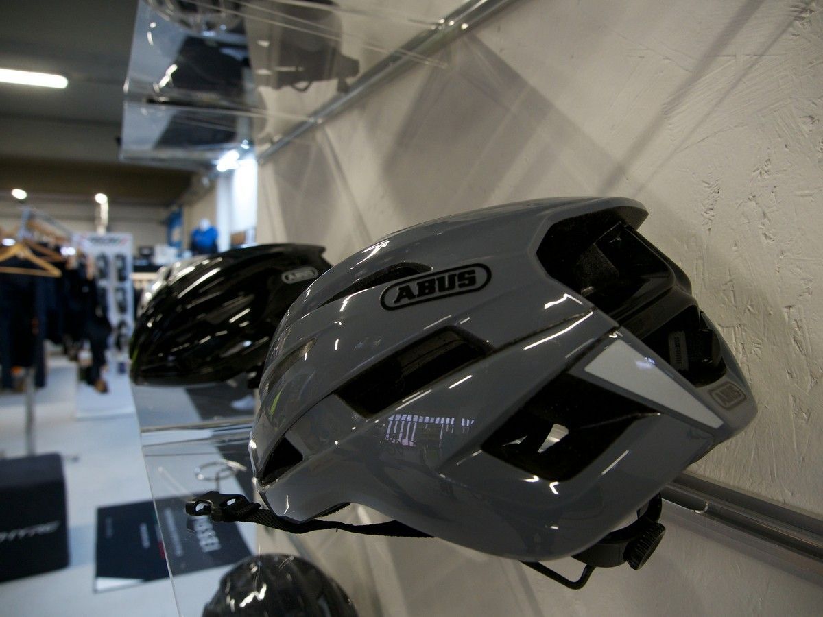 Un casco da bicicletta ABUS grigio esposto su uno scaffale in un negozio, con un altro casco nero visibile sullo sfondo.