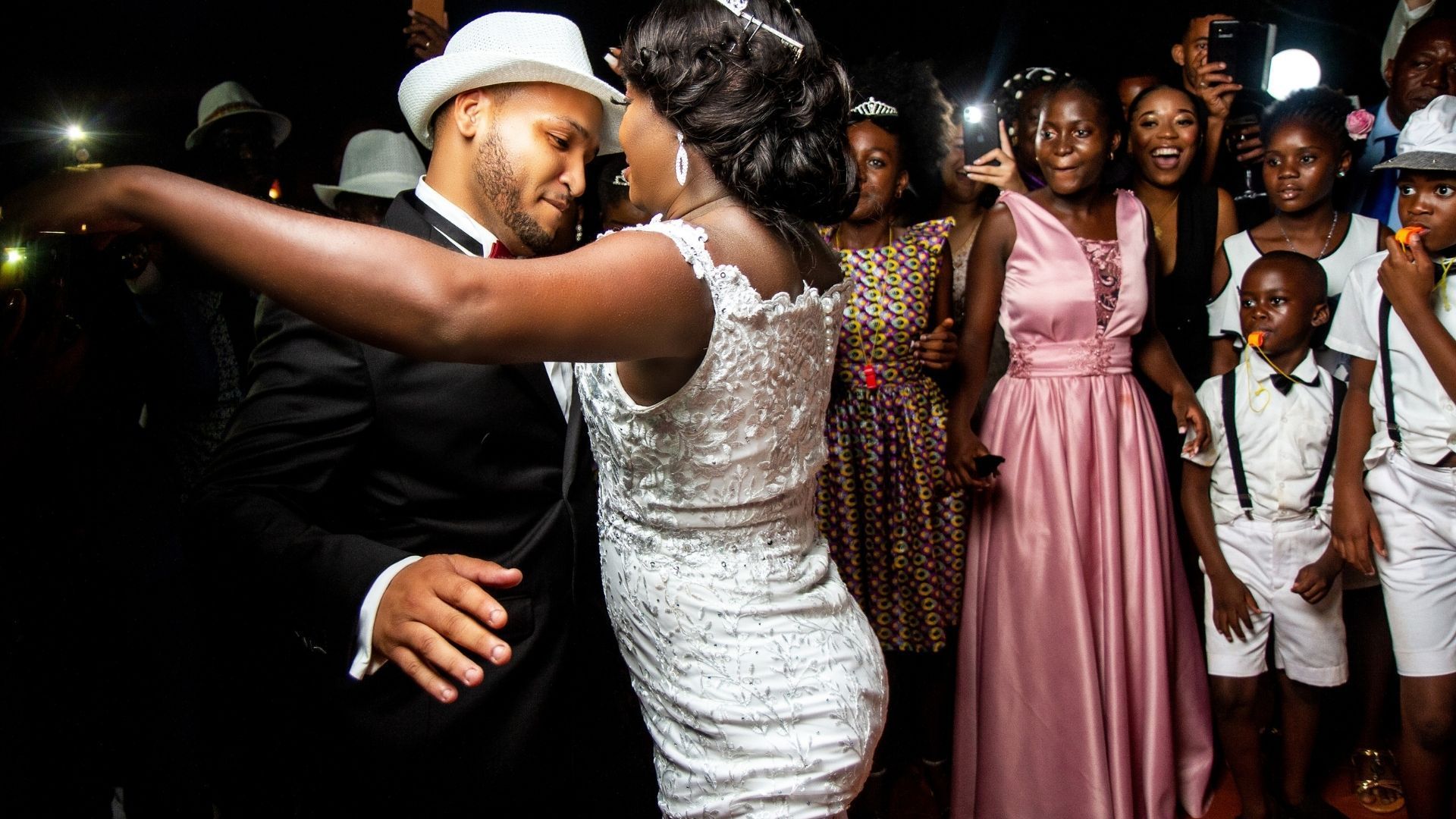 Couple dancing at a party surrounded by guests taking photos. Formal attire, celebratory atmosphere.