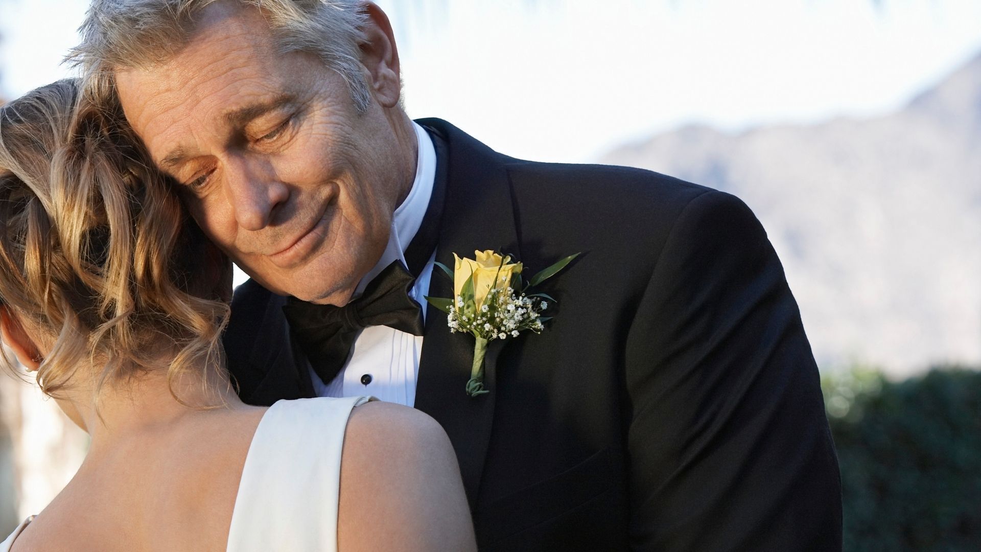 Father and daughter embrace at a wedding. Father smiles, wearing a tux with a yellow rose.