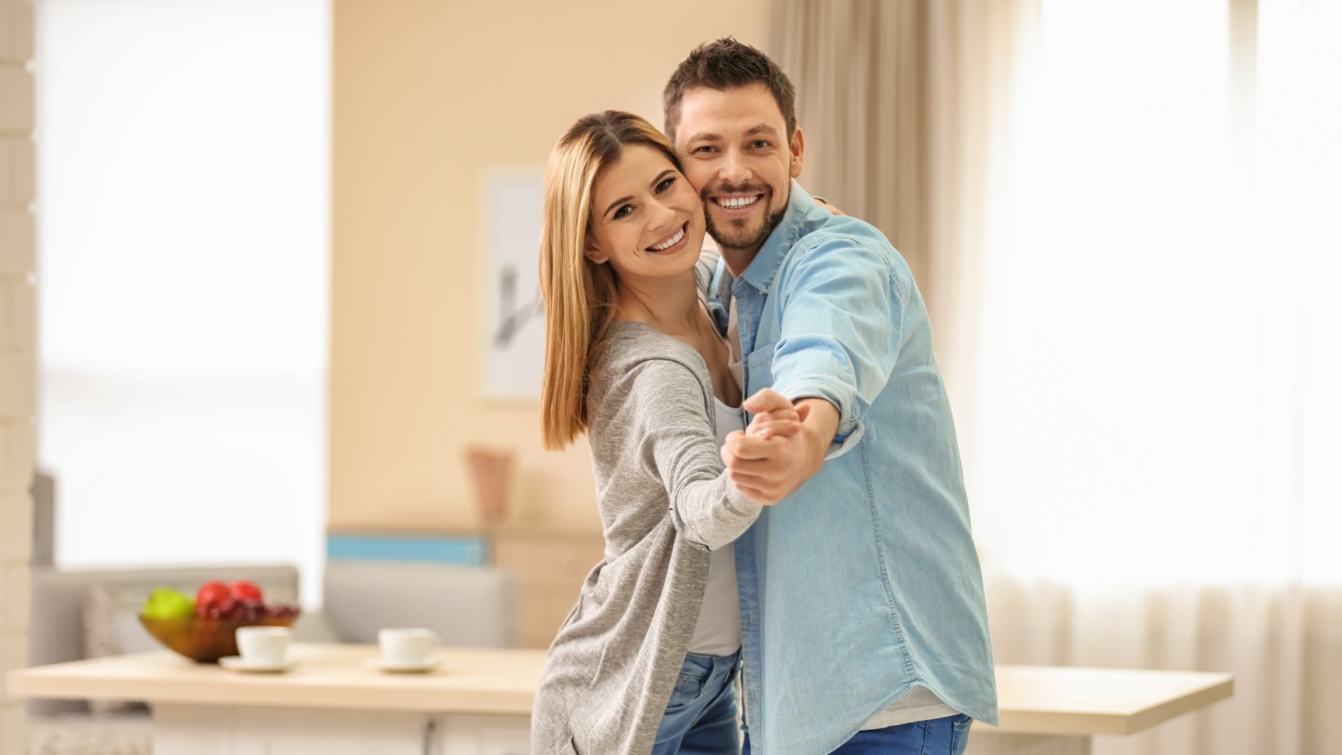 Couple dancing in a well-lit home, smiling at the camera. The woman wears a sweater; the man, a blue shirt.