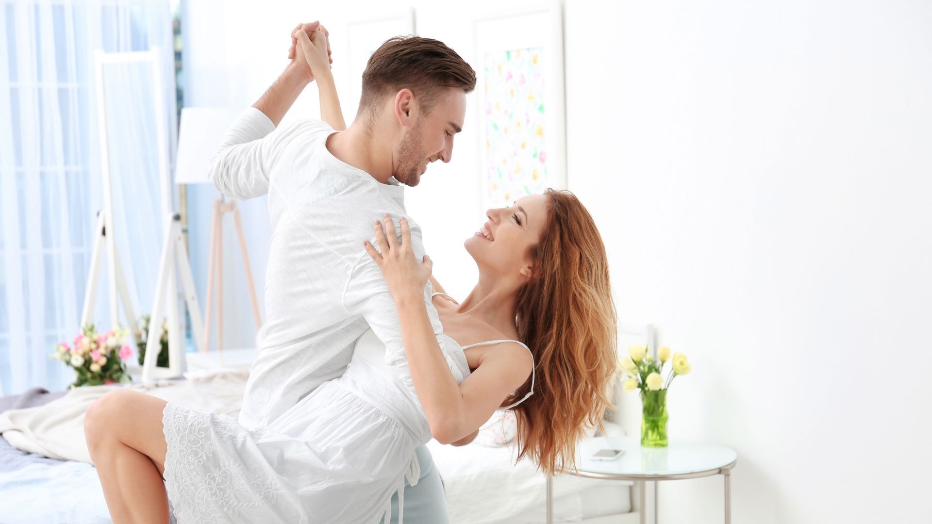 Couple dancing and smiling in a bright bedroom, man in white holding woman in a white dress, happy expressions.