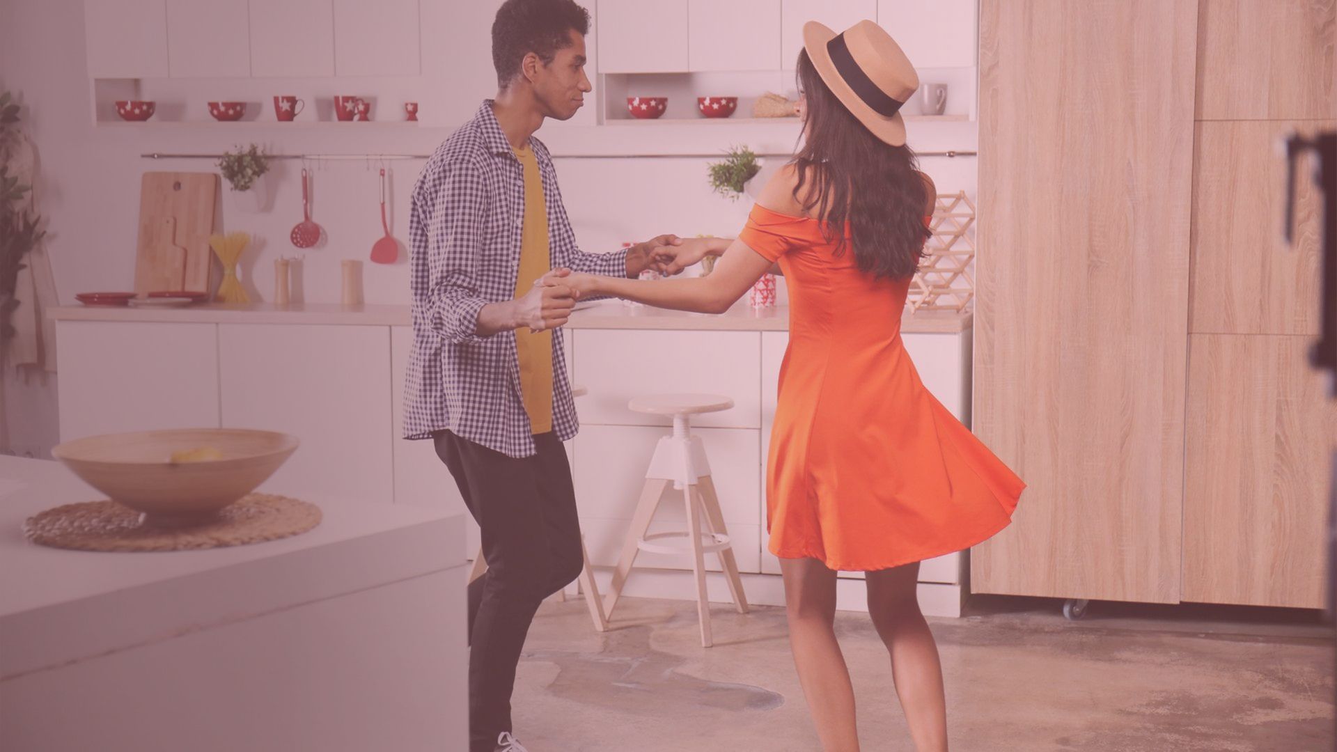 Couple dancing in a kitchen; the woman wears an orange dress, hat; man in a patterned shirt, black pants.