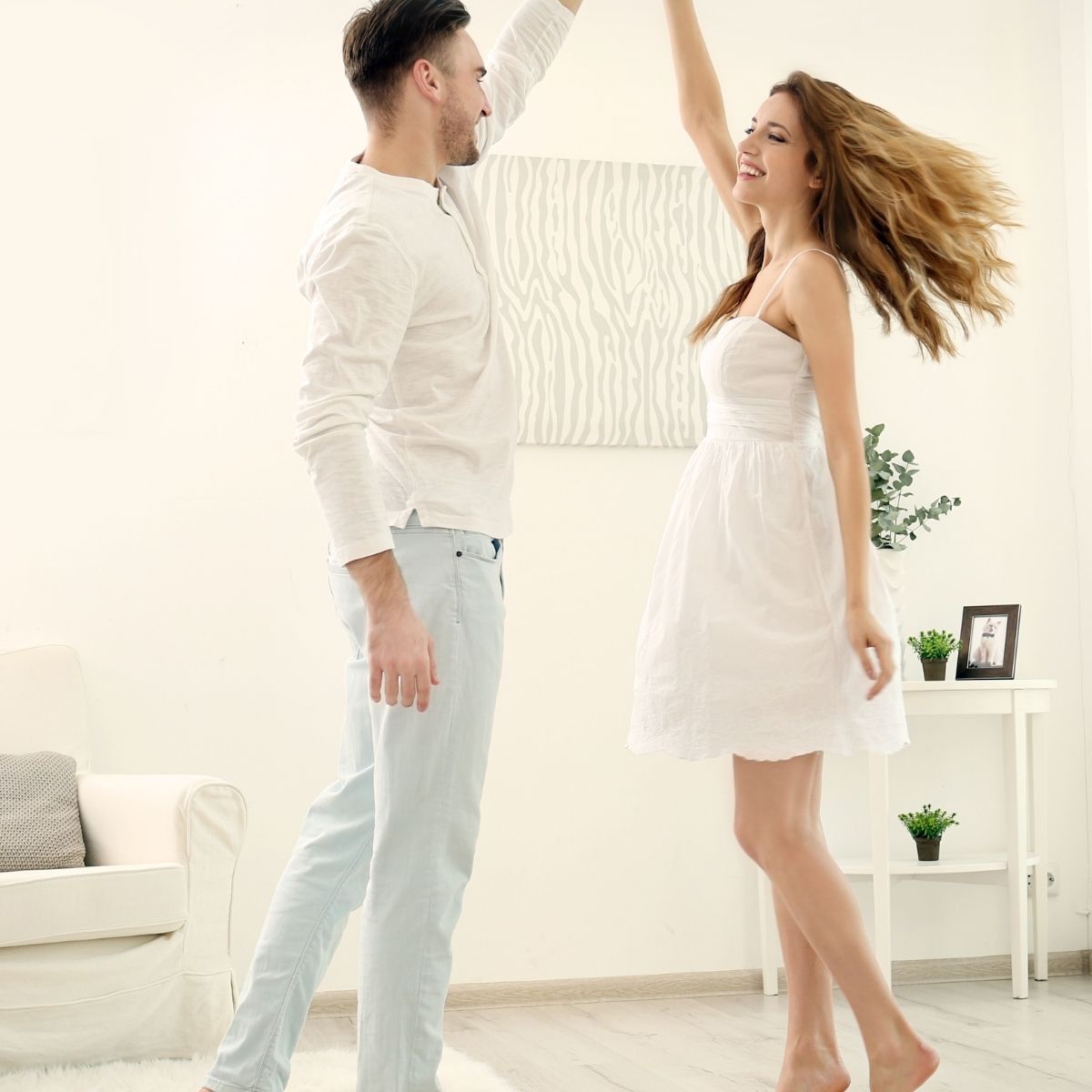 Couple dances joyfully in a bright room. Woman in white dress twirls; man in light clothes smiles, arm raised.