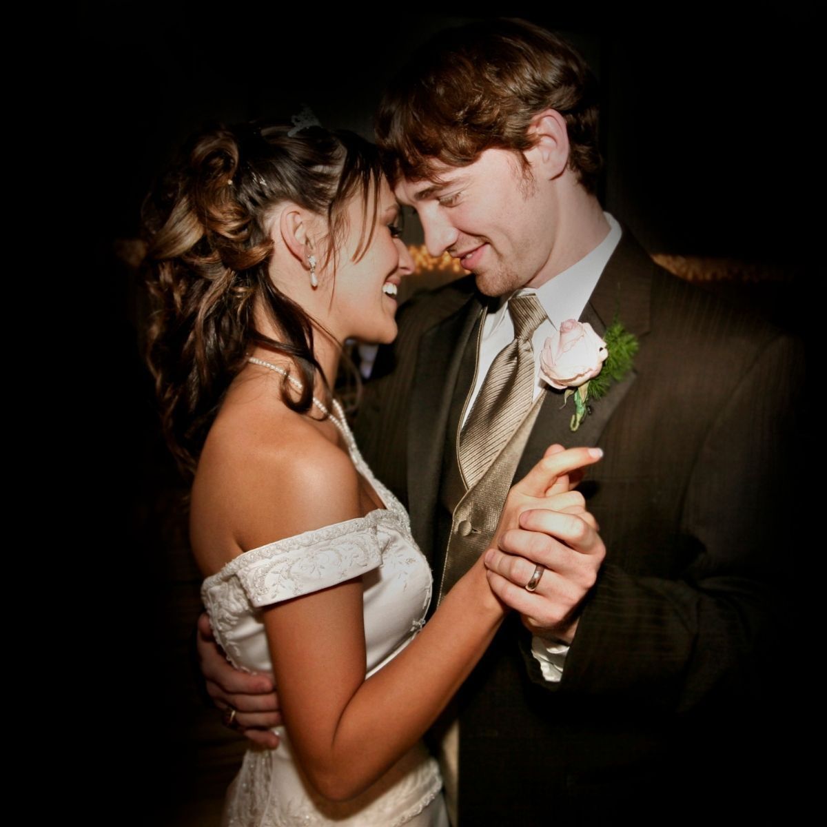 Bride and groom in formal wear dancing, smiling, and gazing at each other in a dimly lit space.