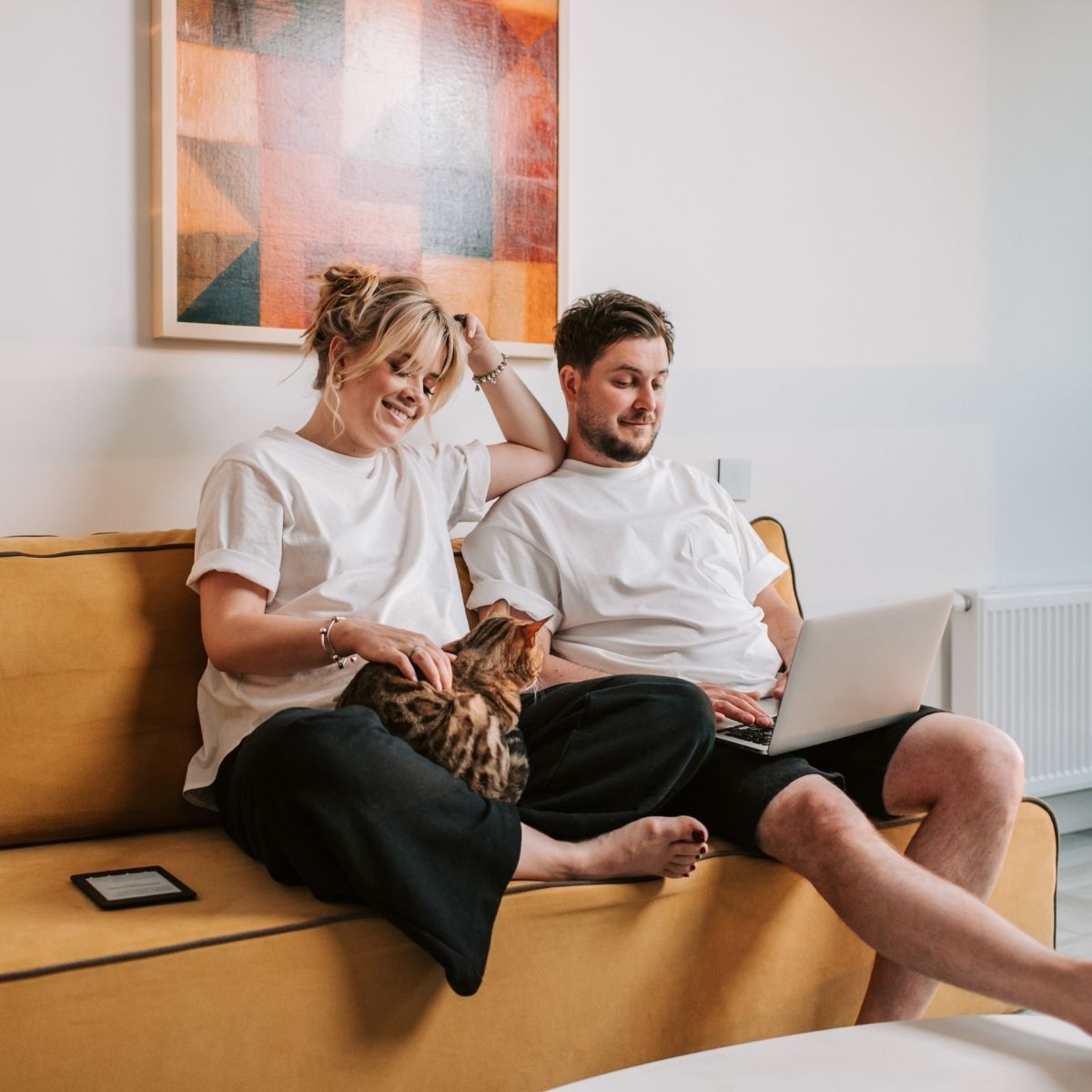 Couple on yellow couch, cat on lap, man using laptop, woman smiling, artwork on wall.