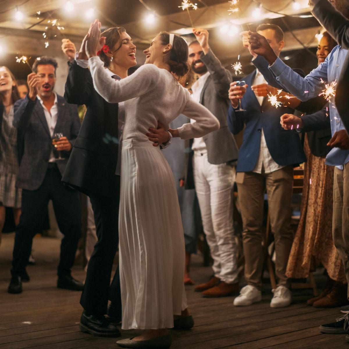 Couple dances at a wedding, surrounded by cheering guests holding sparklers and drinks under string lights.