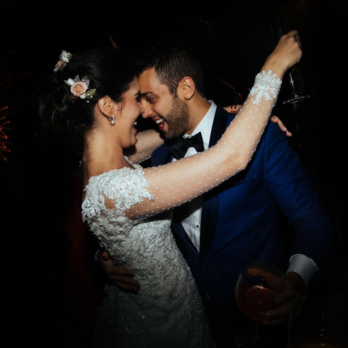Bride and groom dancing close, laughing. She's in white lace, he's in a blue tuxedo. Dark setting.