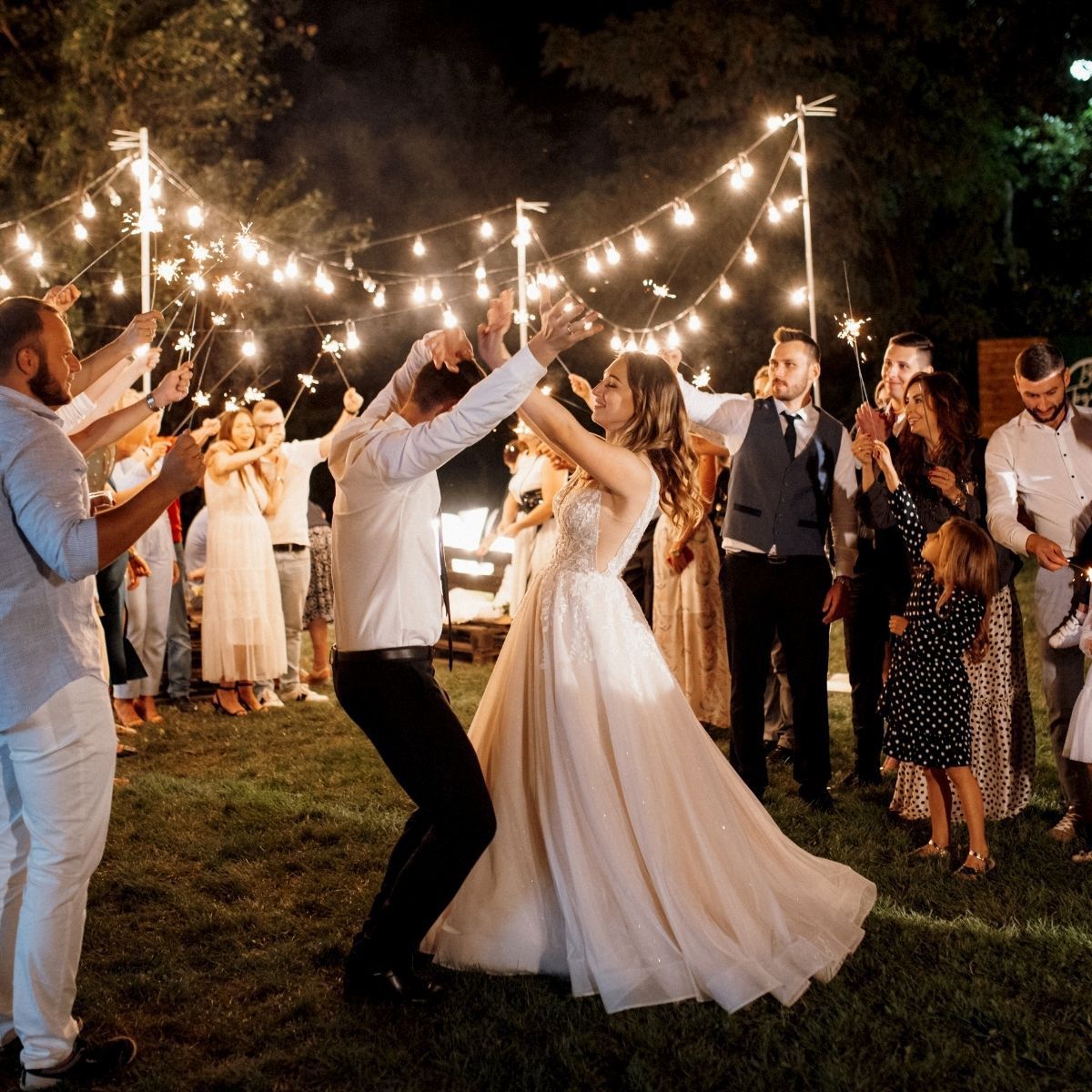 Bride and groom dancing outdoors at night, surrounded by guests holding sparklers and string lights.