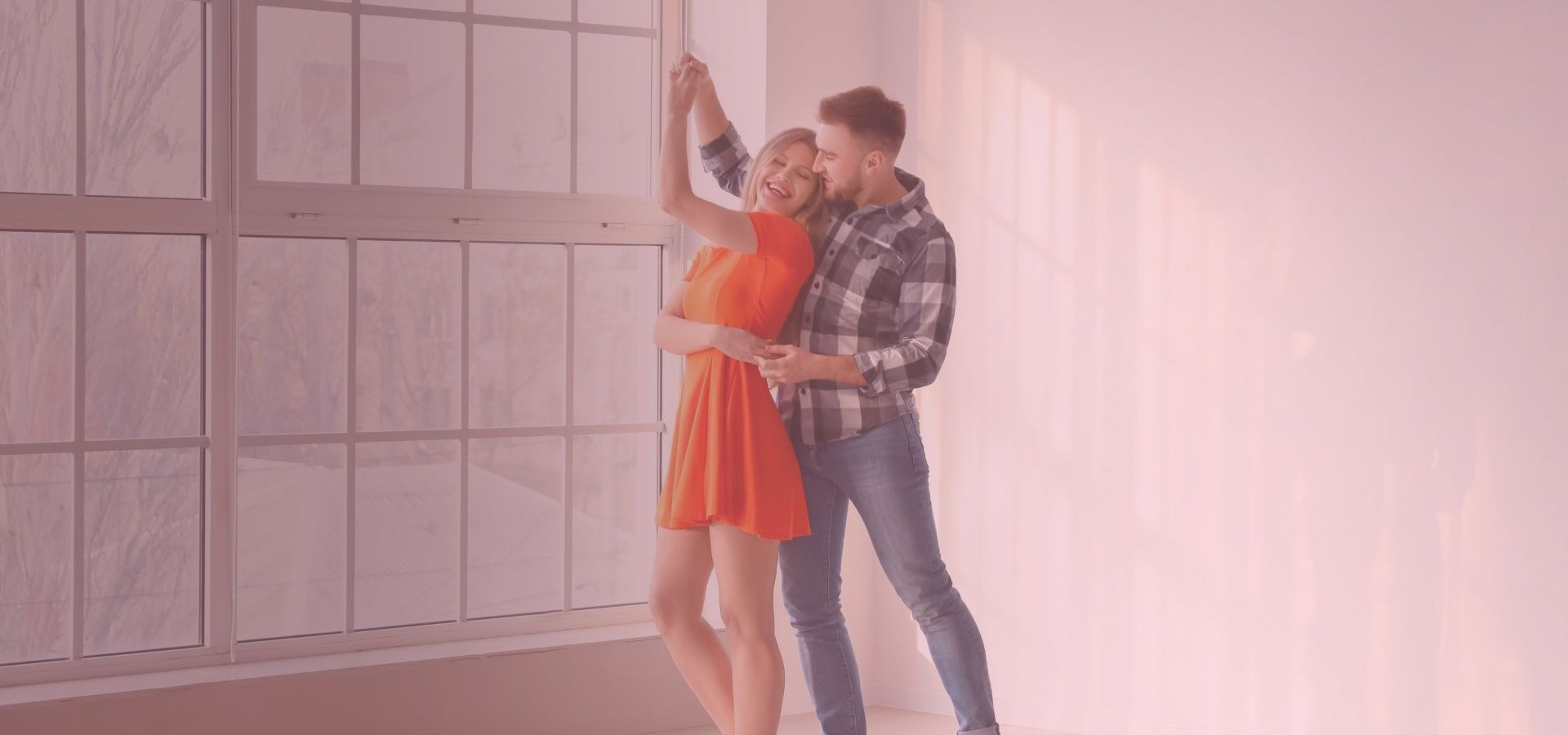 Couple dances indoors, woman in orange dress smiles, man in jeans and plaid shirt.