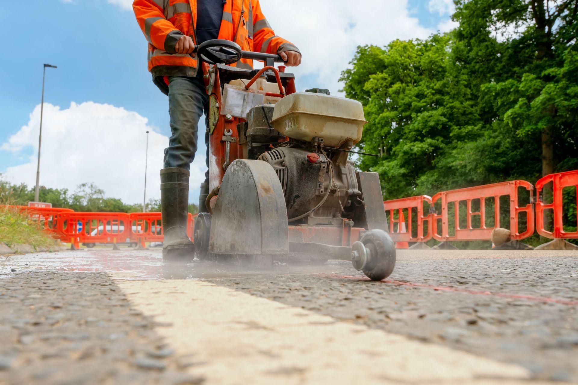 Worker operating road saw, cutting into asphalt road surface. Orange safety barriers and vest.