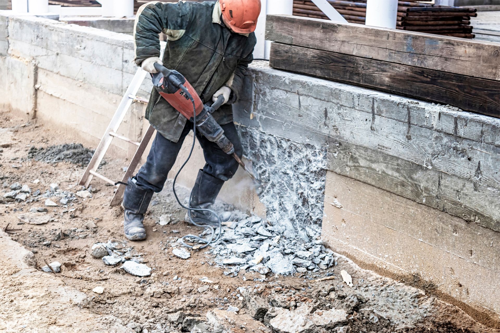 Construction worker in orange hard hat uses a jackhammer to break concrete wall outdoors.
