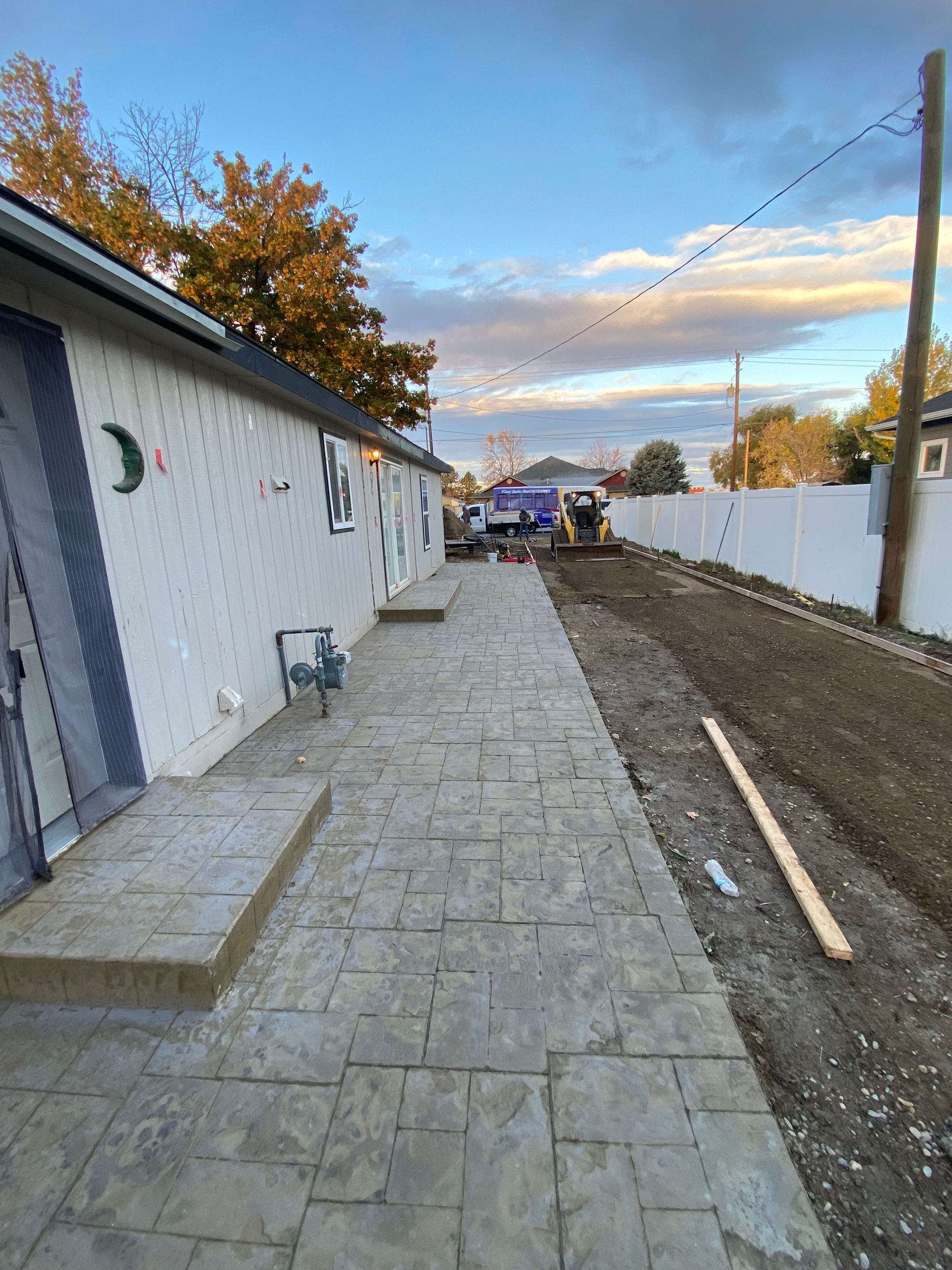 A long stone walkway beside a white building and a dirt path with construction equipment.