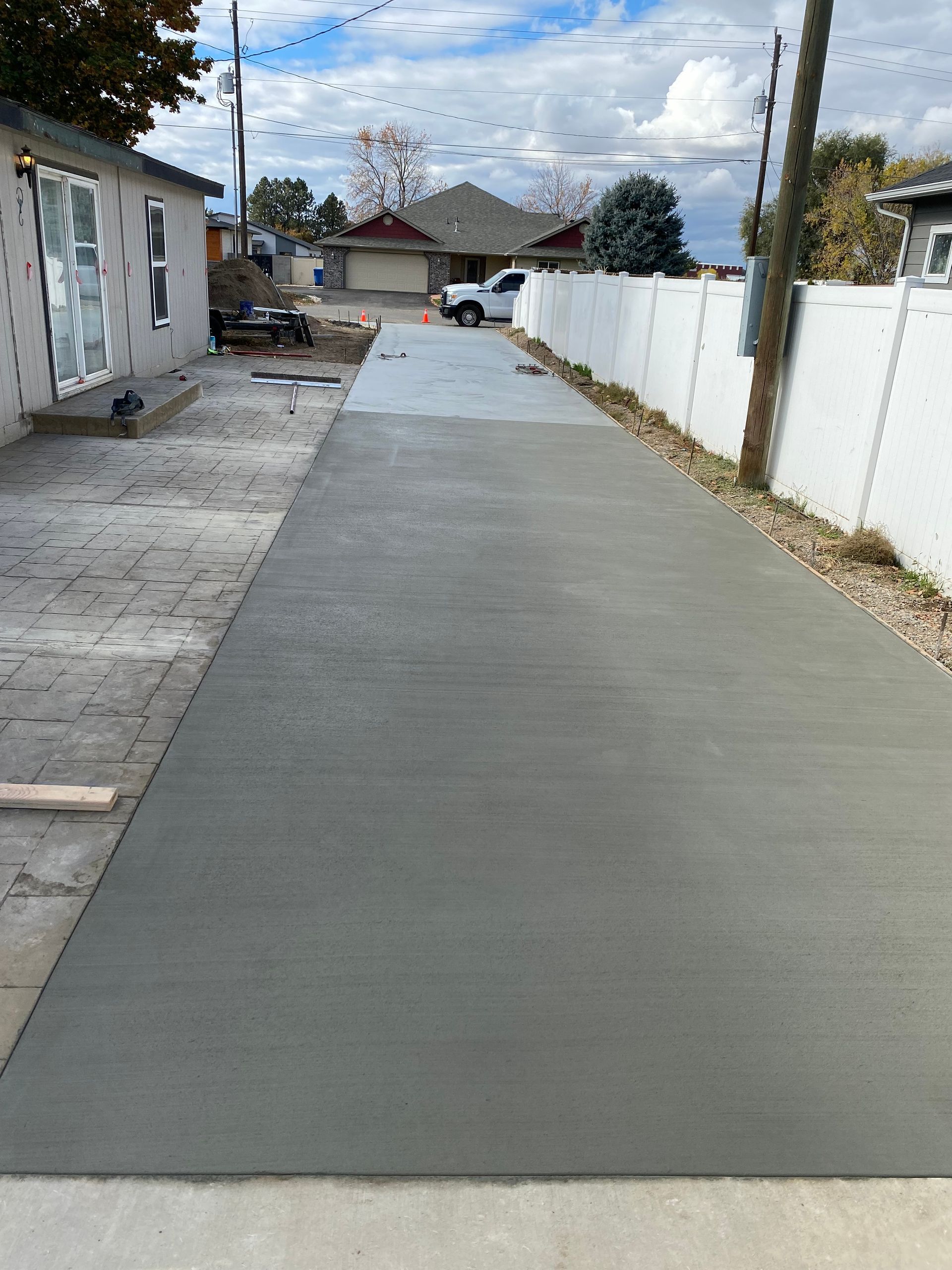 Freshly poured concrete driveway. Gray surface, bordered by a white fence and a building.