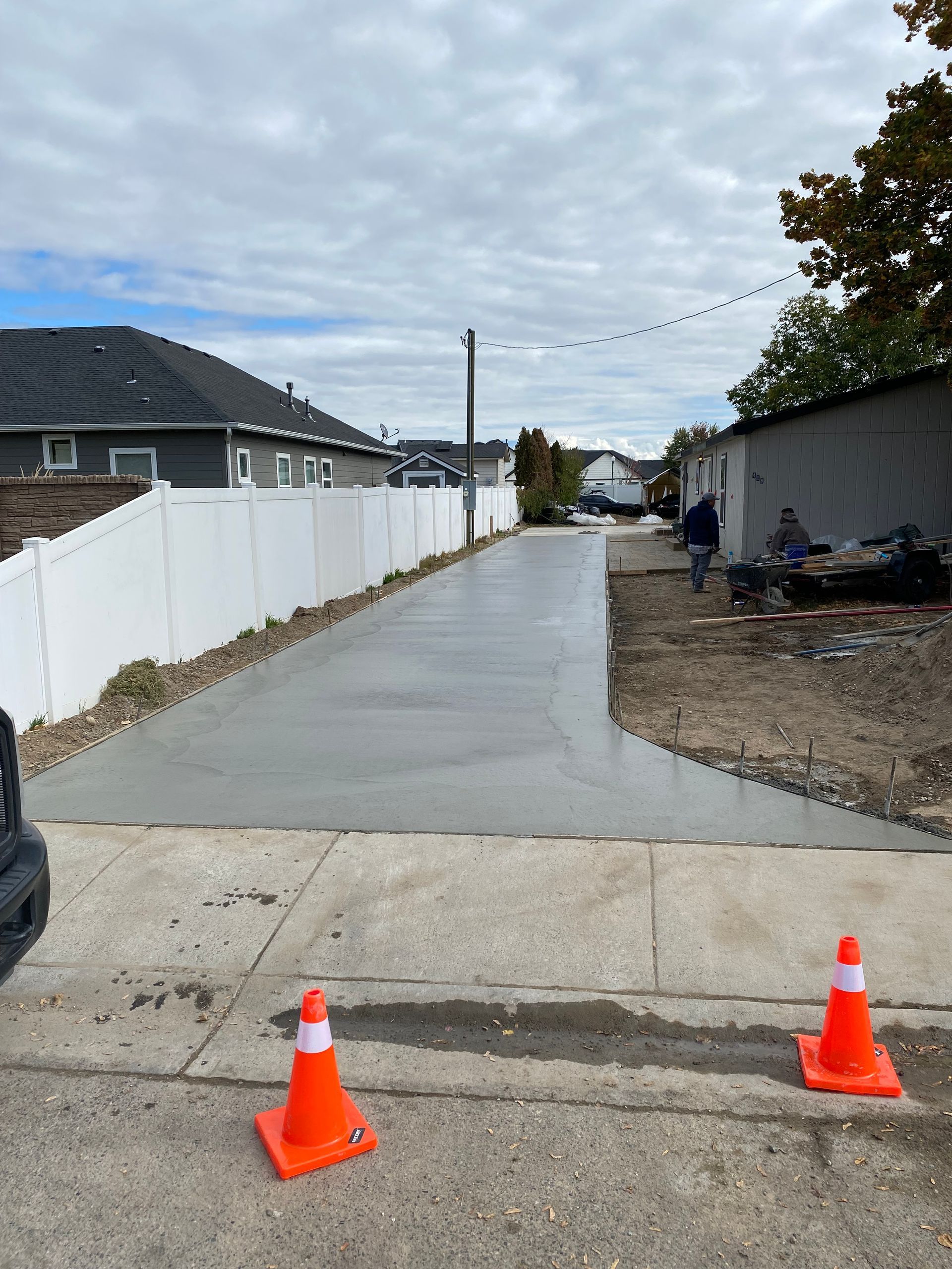 Newly poured concrete driveway extending between white fence and building; orange traffic cones in foreground.