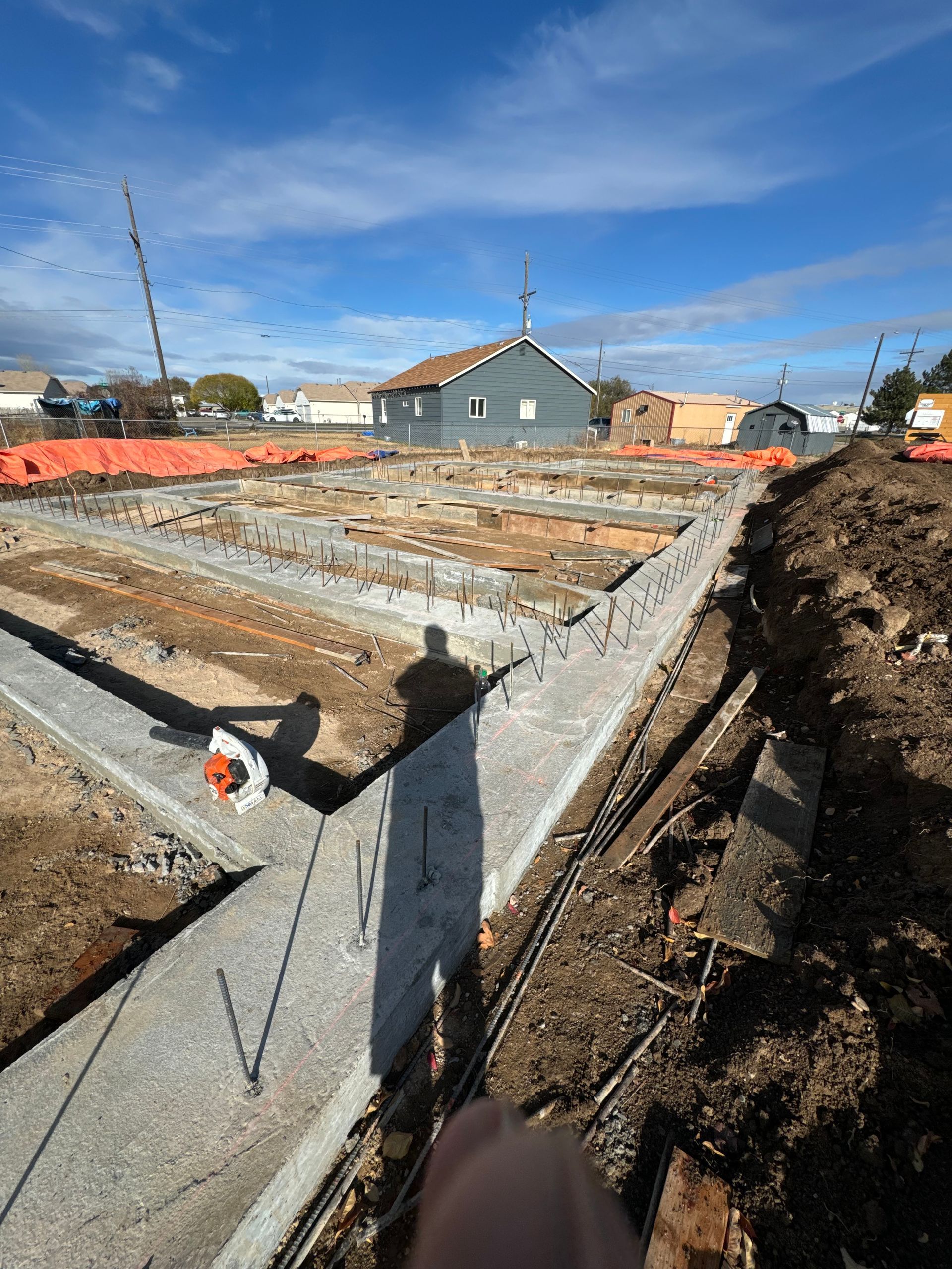 Construction site with concrete foundation walls; house in the background under a blue sky.