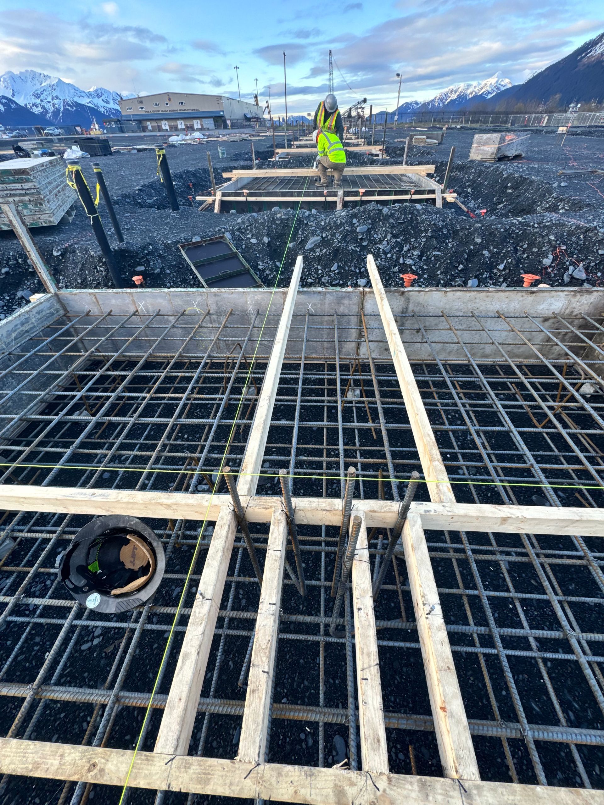 Construction site: Rebar grid with wooden forms, worker in high-vis vest, mountains in the background.