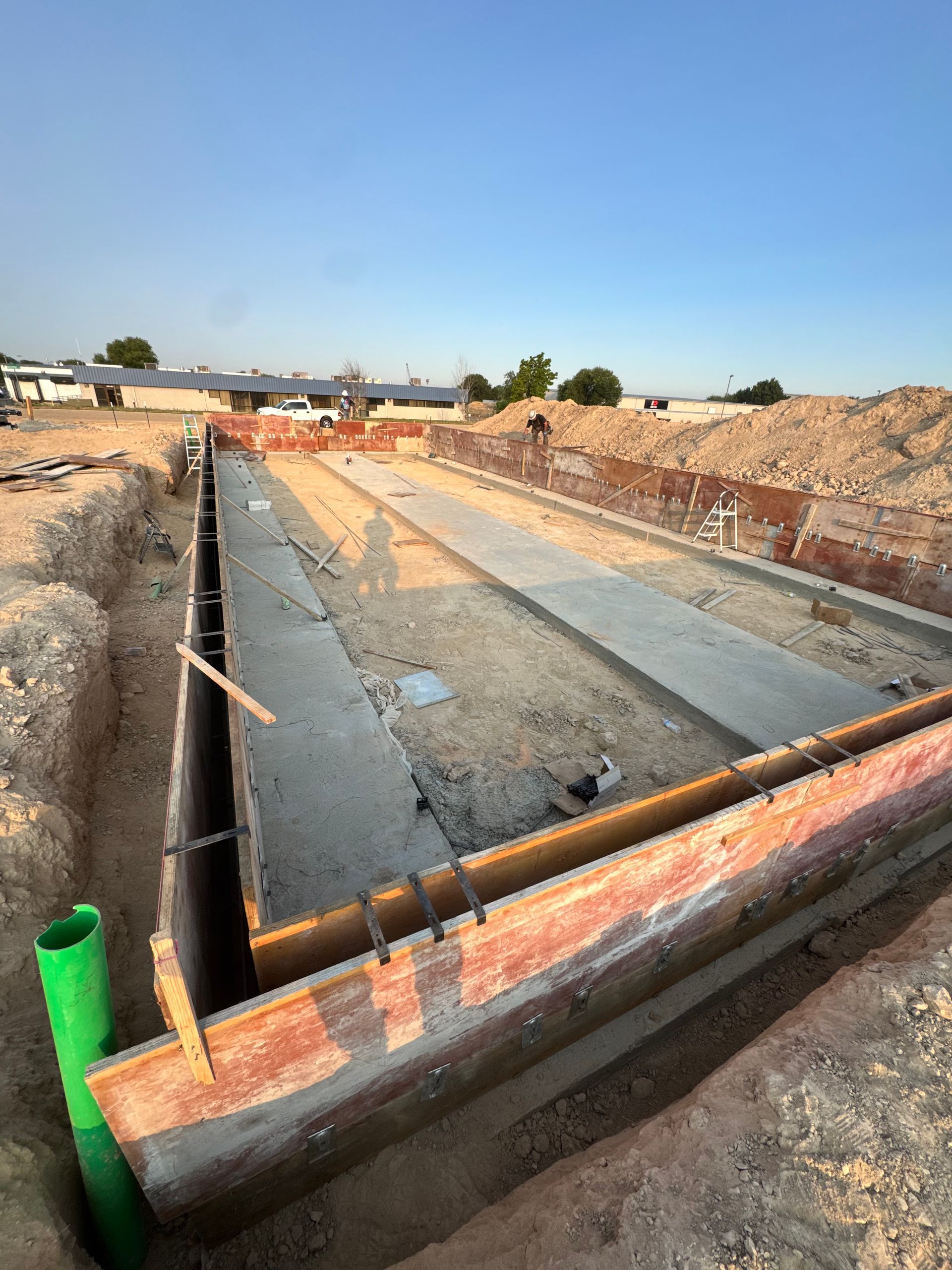 Construction site with rectangular concrete forms in a trench, orange framing, dirt, and a blue sky.