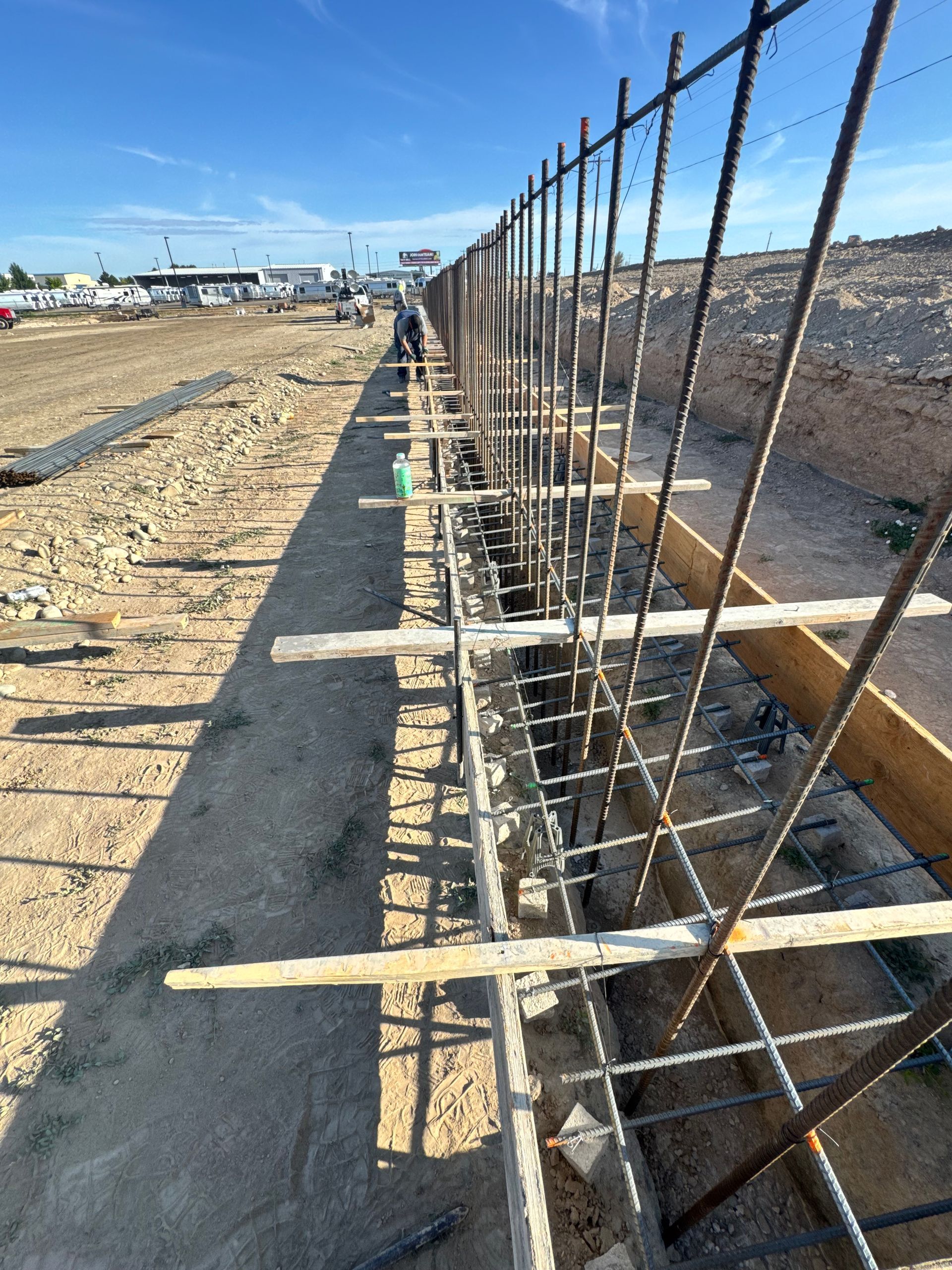 Construction of a tall wall with vertical and horizontal rebar, wooden supports, and a worker in the distance.