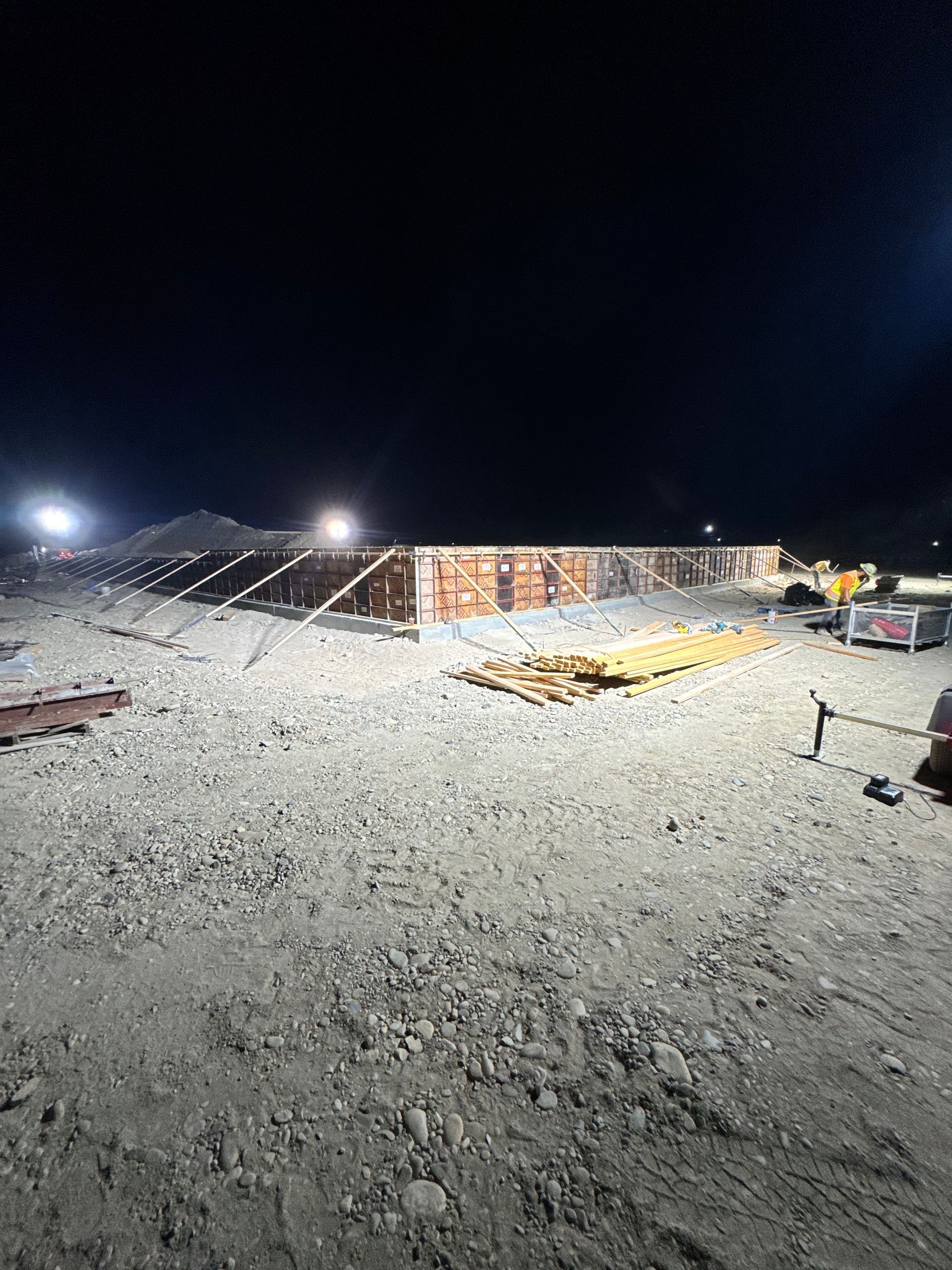 Construction site at night, building materials and machinery illuminated against a dark sky.