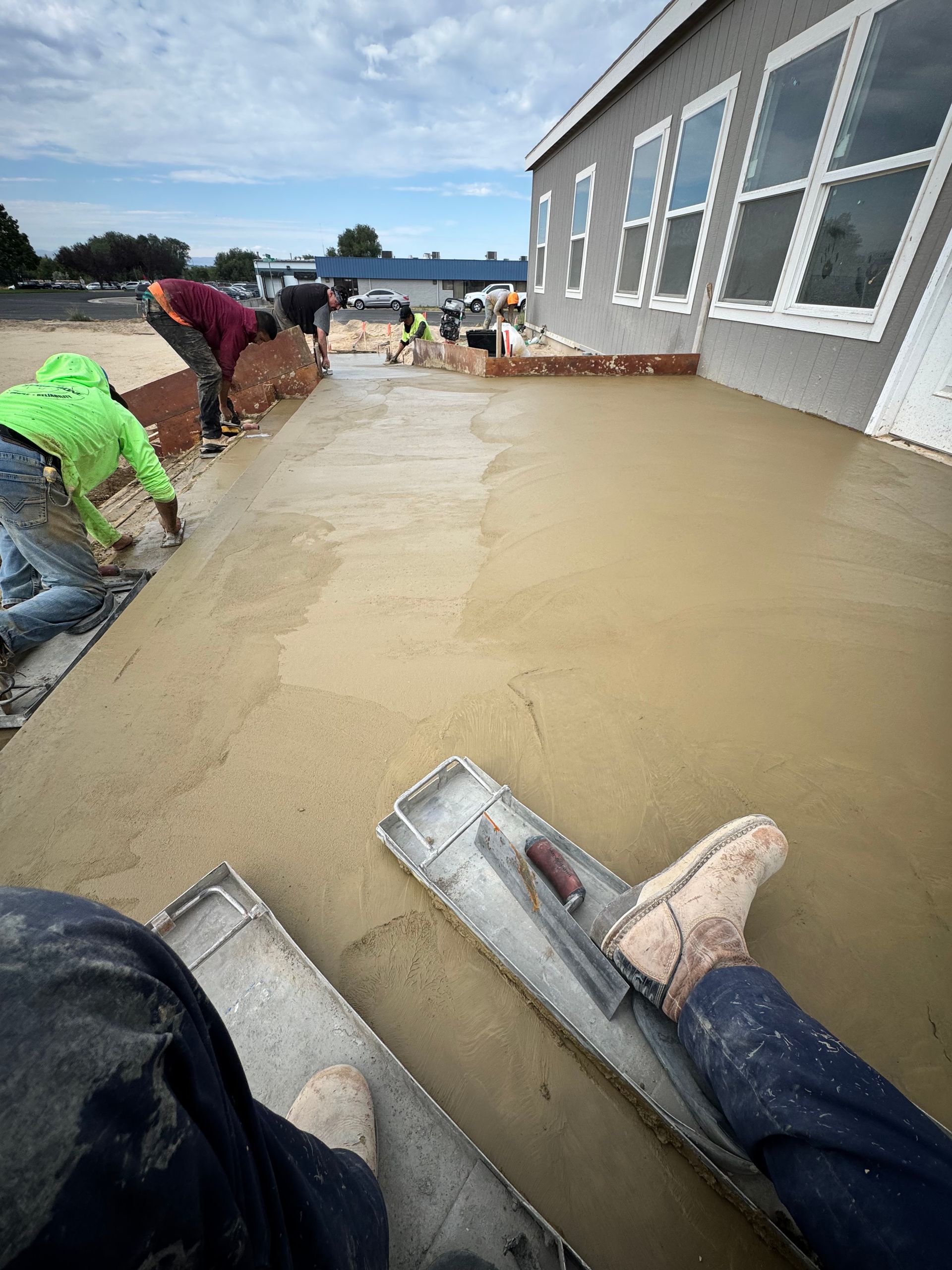 Workers smoothing wet concrete on a sidewalk with building in the background on a cloudy day.