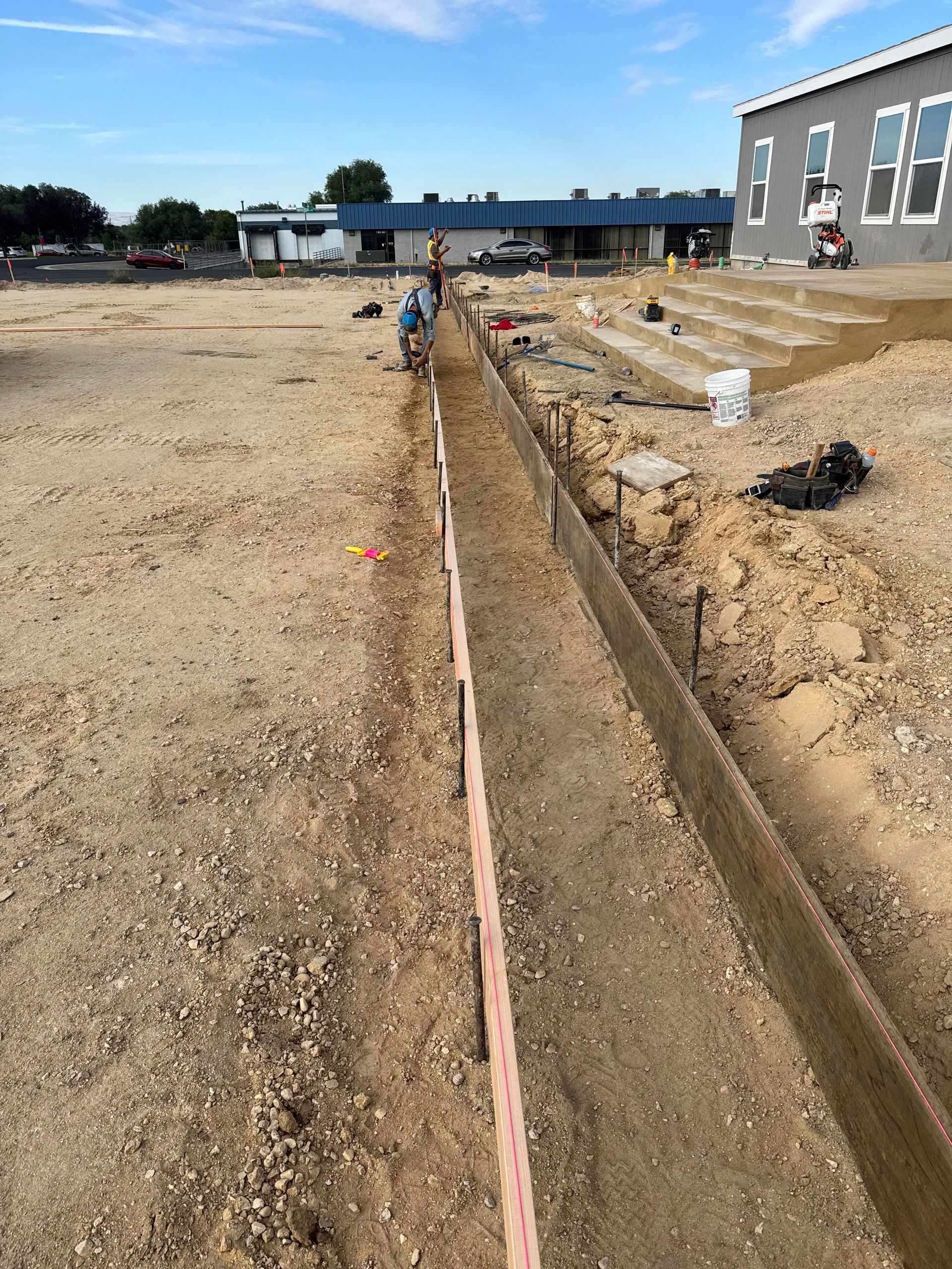 Construction site: trench with wood formwork, tools, workers, and building in the background.