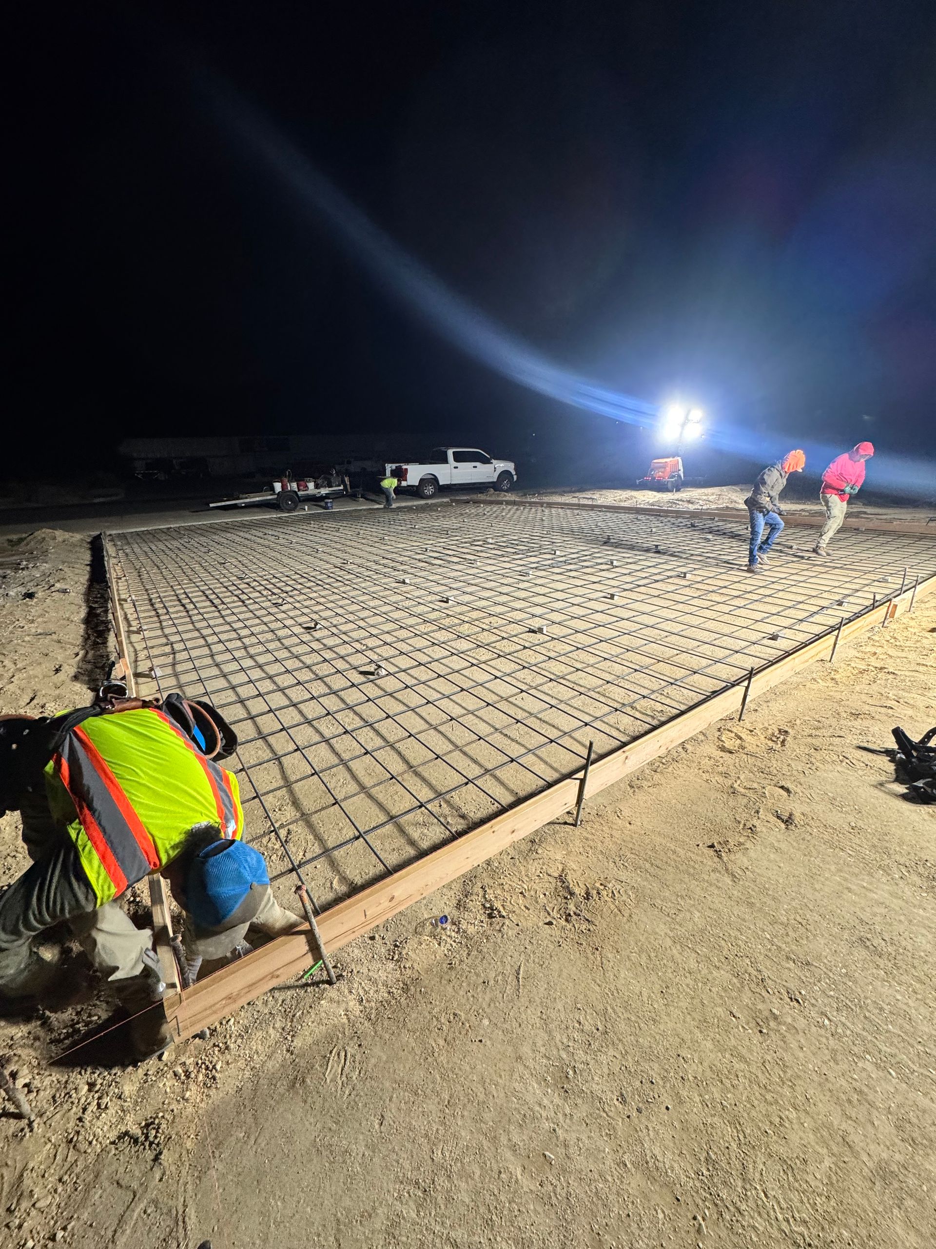 Construction workers laying rebar mesh for a concrete slab at night. Illuminated by bright lights.
