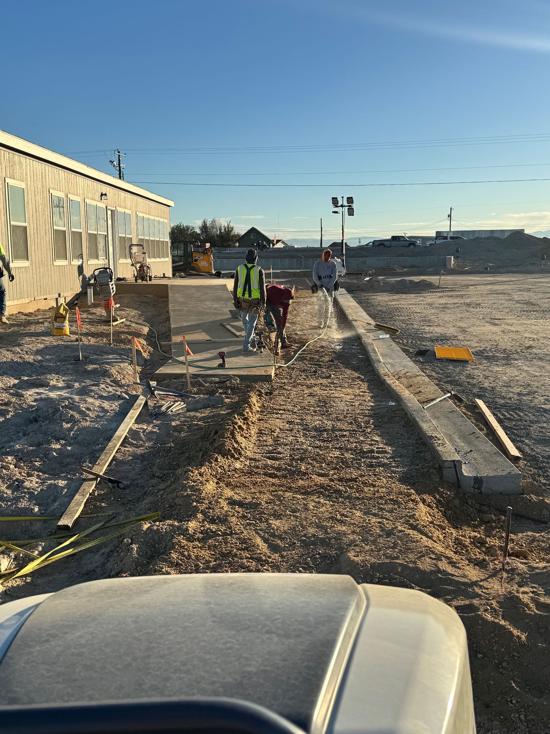 Construction site: workers near a low building, using tools on a dirt path with concrete edging.