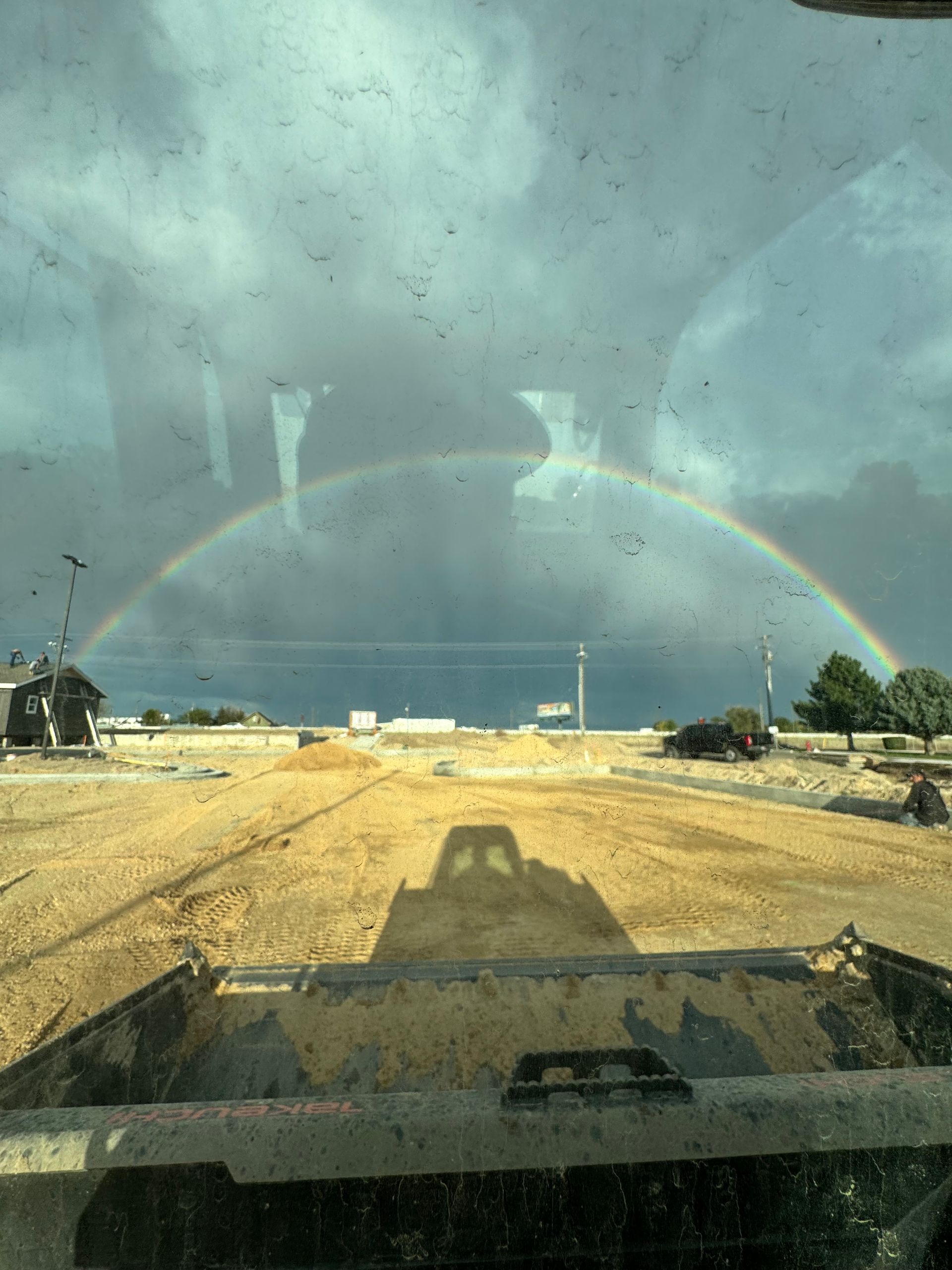 Rainbow arches over construction site; loader shadow visible. Cloudy sky.