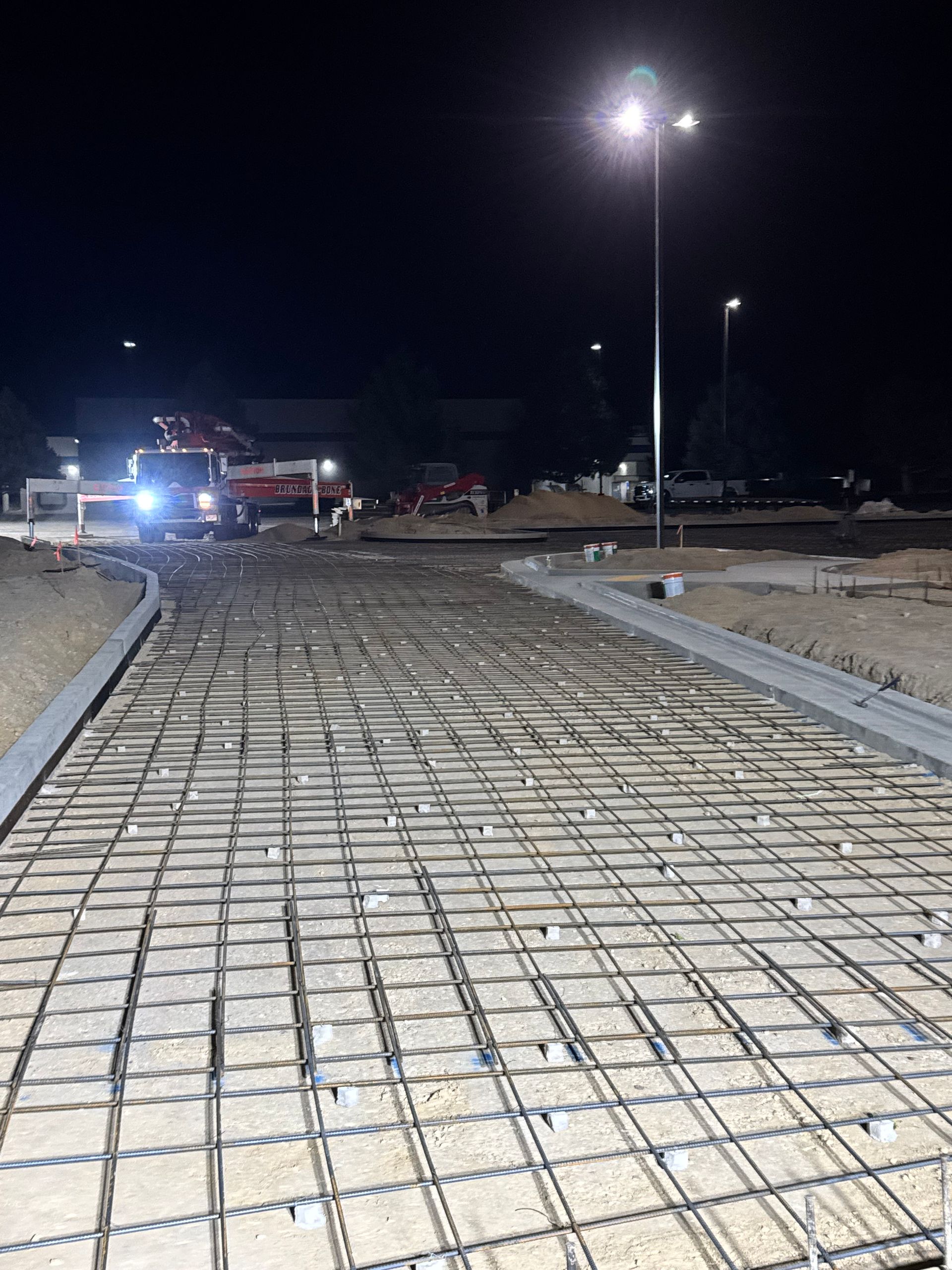 Night construction site with rebar grid laid for road paving. Streetlights illuminate the scene.