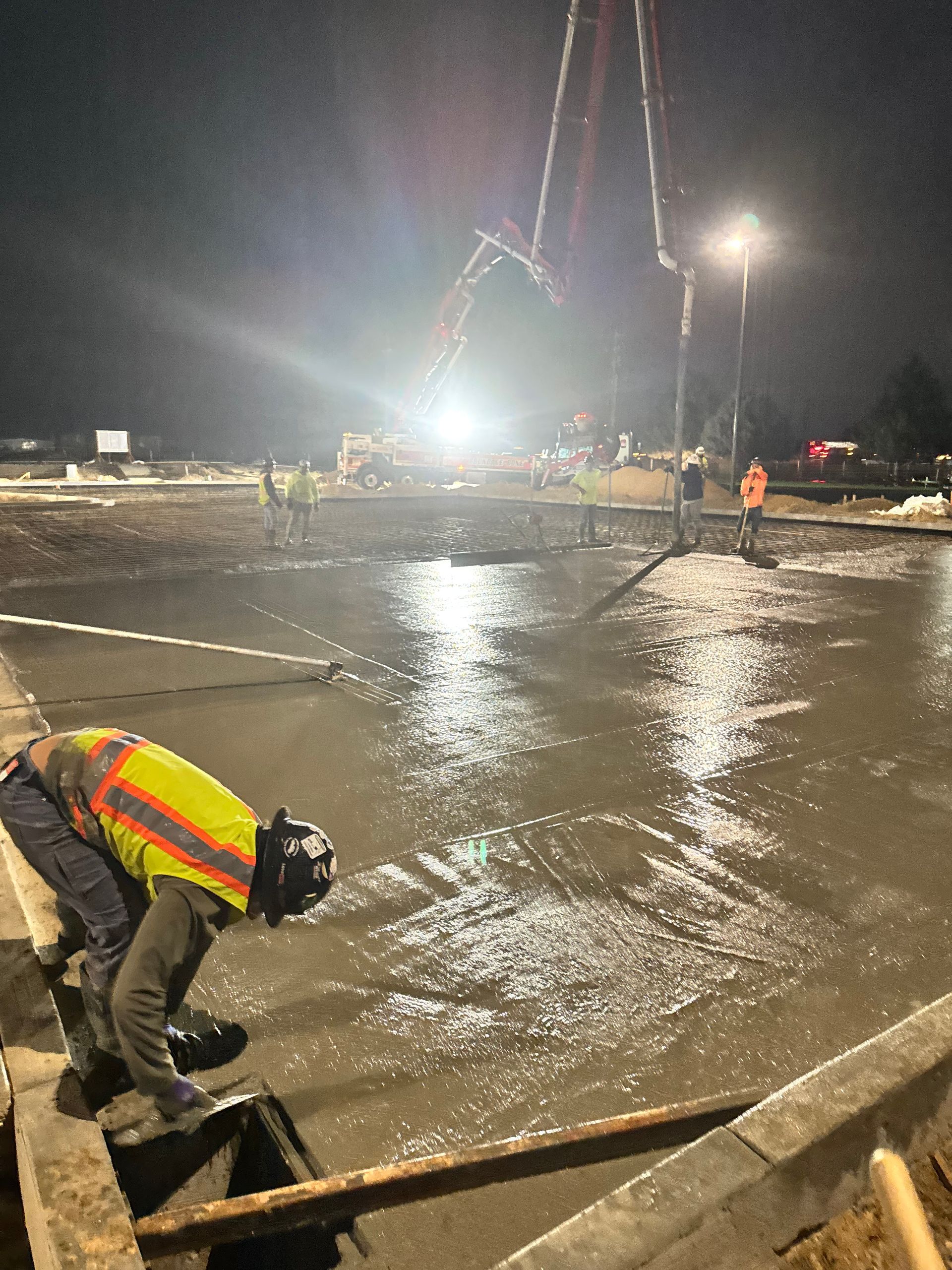 Construction worker smoothing wet concrete at night. A concrete pump and bright lights are visible.