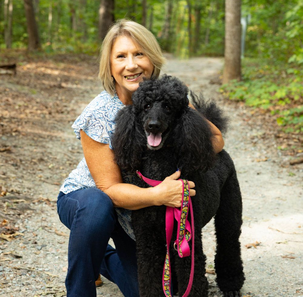 Woman kneeling, hugging a black poodle on a wooded path. Both smiling.