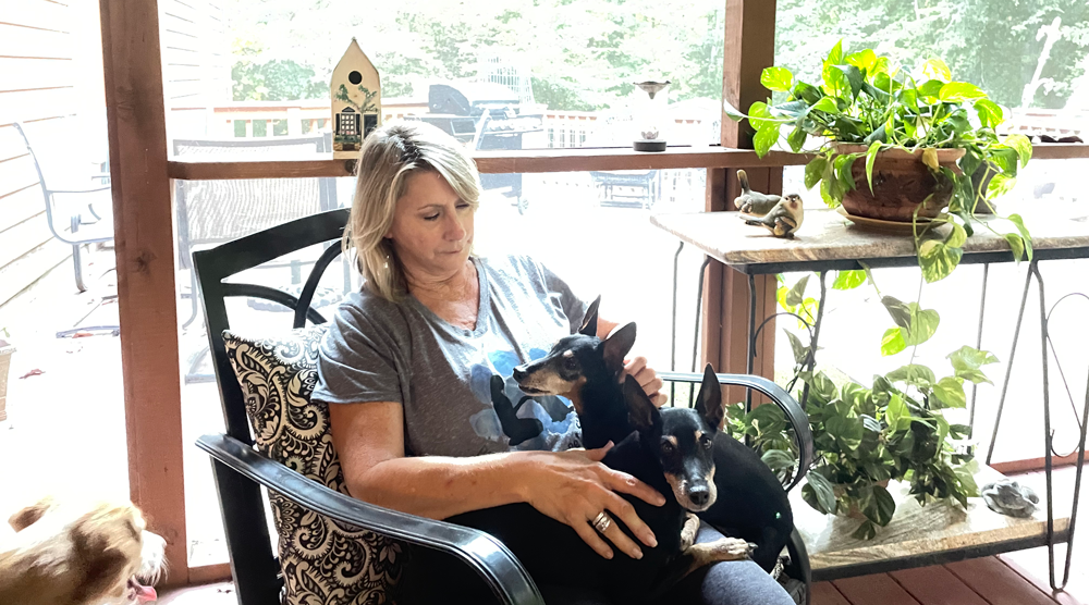 Woman petting two small black dogs on a porch, with plants and a birdhouse in the background.