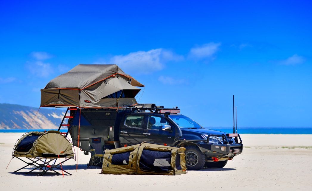 Black truck with rooftop tent parked on a sandy beach under a blue sky, camping gear alongside. — TJM Albury Pty Ltd in South Albury, NSW