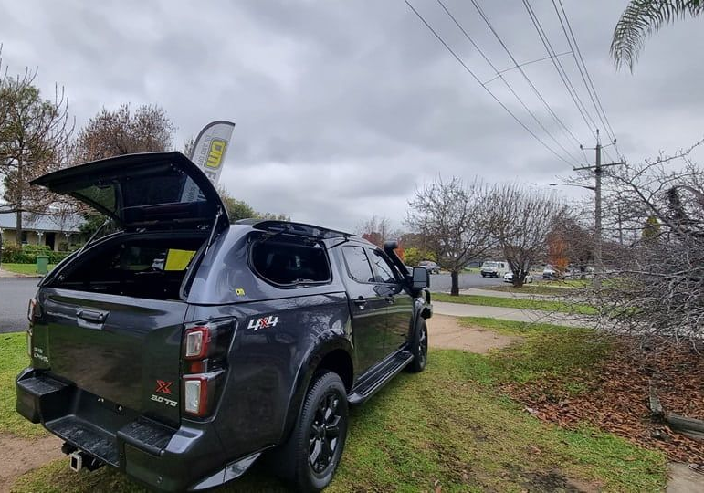 Black pickup truck with camper top parked on grass next to a residential street under an overcast sky. — TJM Albury Pty Ltd in South Albury, NSW