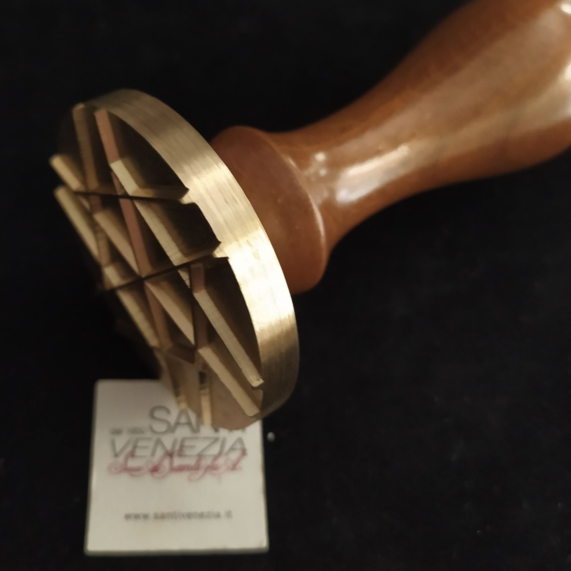 Un timbro in ottone con un motivo a griglia, attaccato a un manico di legno, sospeso sopra un piccolo cartoncino con la scritta