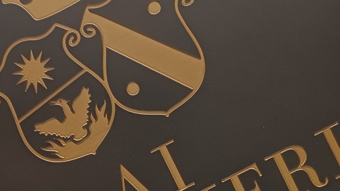 Uno stemma dorato raffigurante un sole e un uccello, su sfondo scuro, con parte della scritta "ALFERI" visibile.