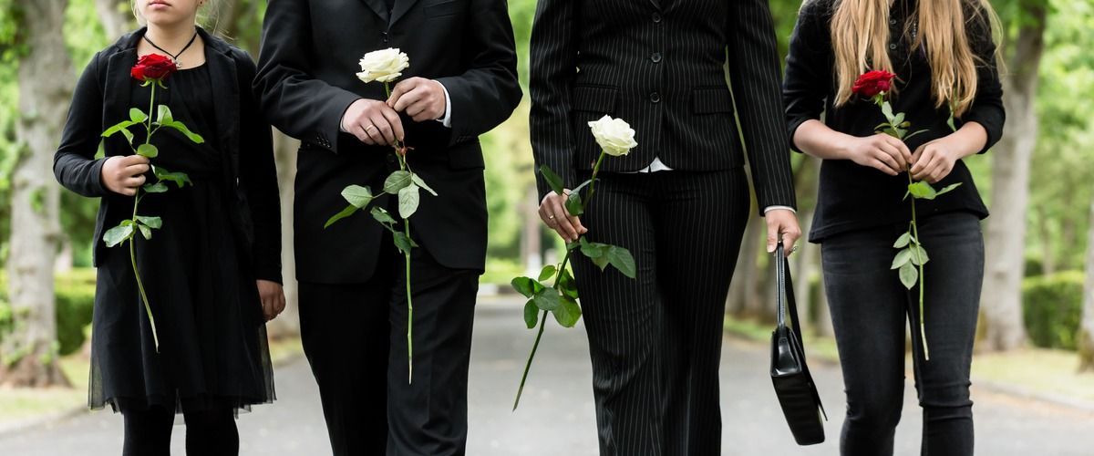 Four people in black clothing holding roses walk down a path lined with trees.