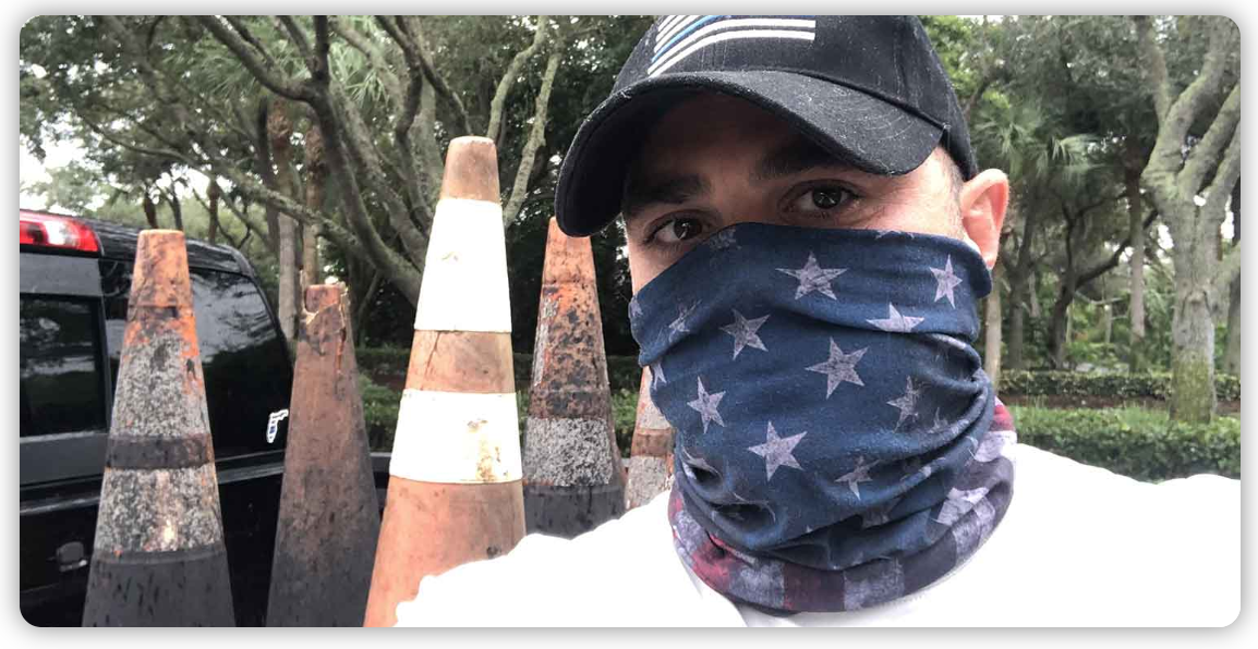 A man wearing a hat and a bandana is standing in front of traffic cones.