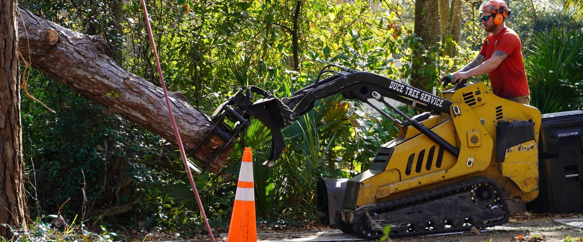 A person in safety gear operates a small yellow skid steer machine to lift a large tree trunk in a wooded area.