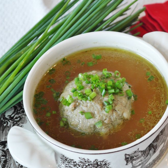 Eine Schüssel Rindssuppe mit einem großen Leberknödel, garniert mit gehackten Frühlingszwiebeln und frischem Schnittlauch daneben.