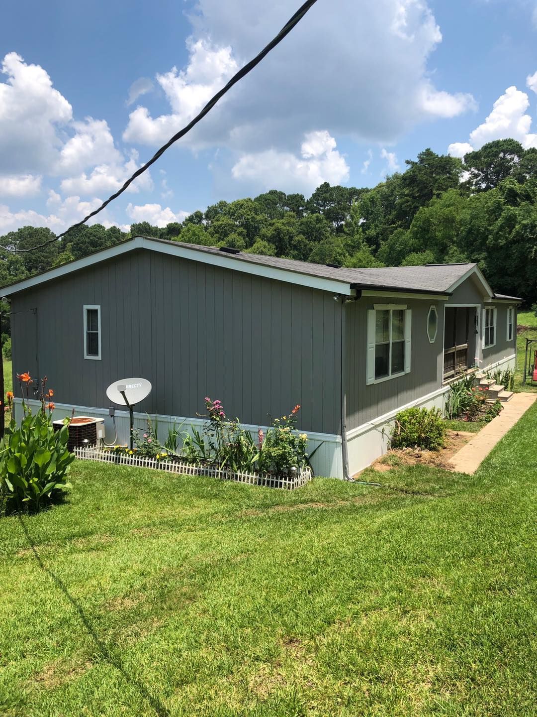 Gray house with white trim and a satellite dish, surrounded by a green lawn and a flower bed. Trees are visible in the background.